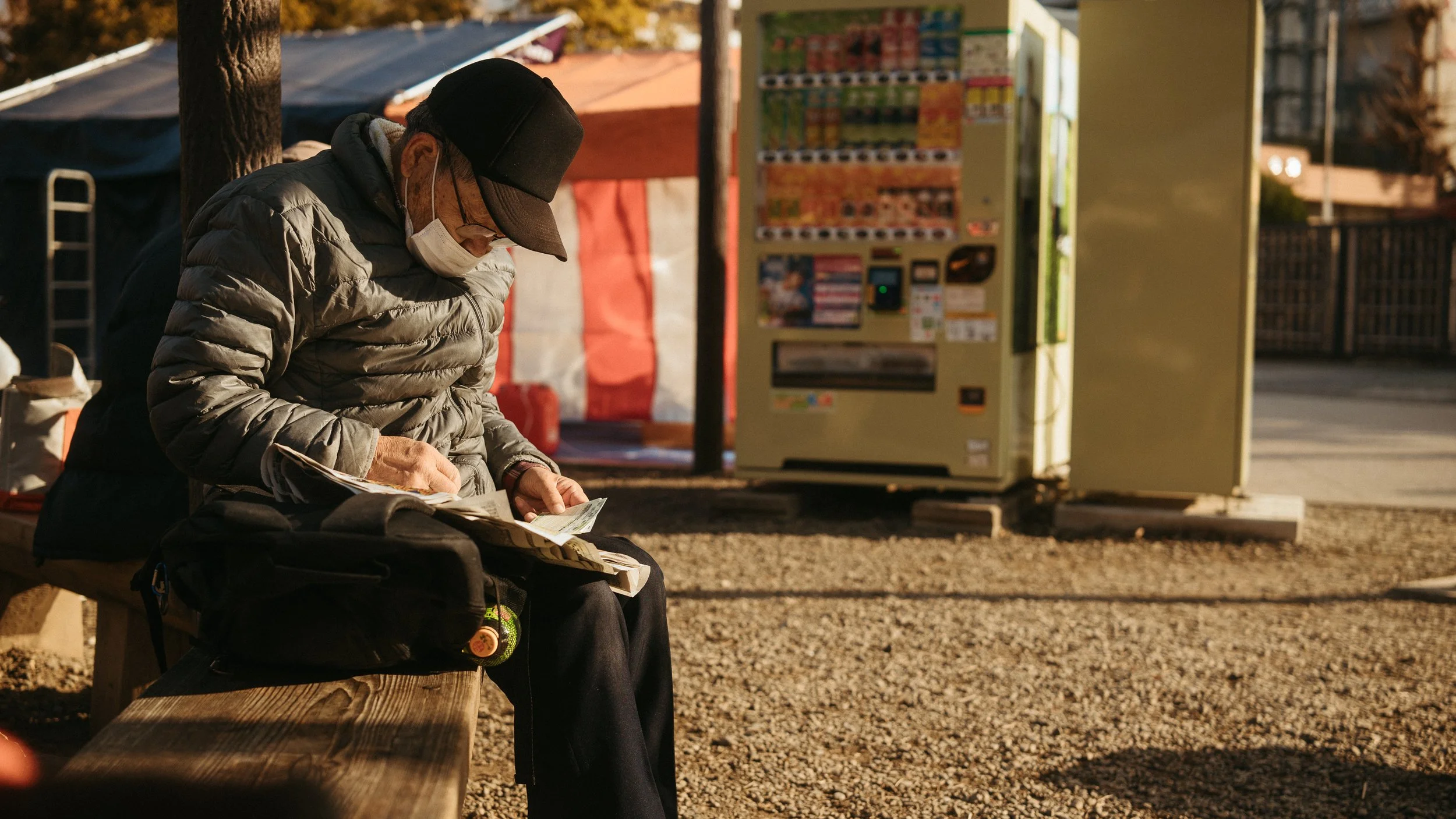 An elderly man with a face mask, glasses, a black cap, and a gray puffy jacket sitting on a wooden bench at a bus stop, looking at papers or a newspaper. Behind him are vending machines, a tent, and trees with autumn foliage.