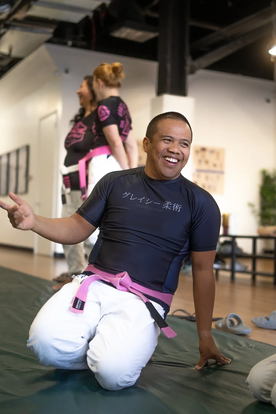 A man in a black martial arts shirt and white gi pants with a pink belt, smiling and kneeling on a martial arts mat, during a martial arts class or training session.