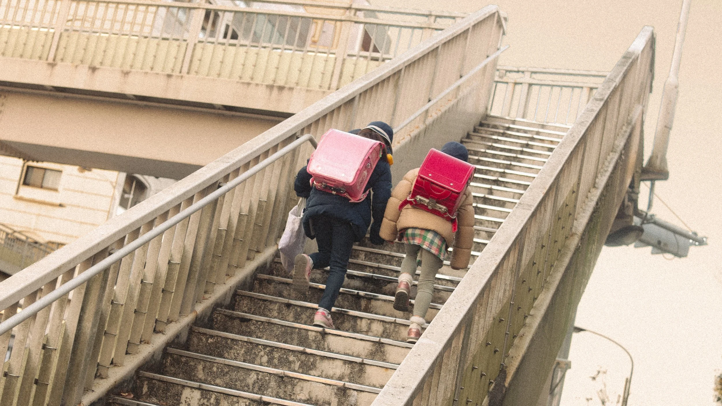 Two children carrying pink and red backpacks walk up an outdoor staircase.