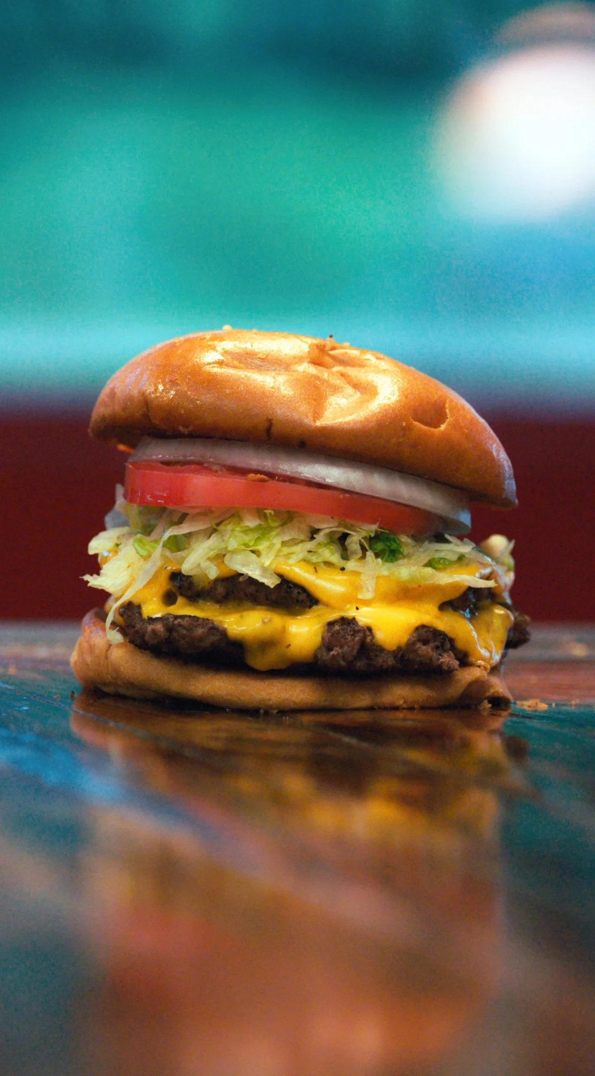Close-up of a cheeseburger with lettuce, tomato, onion, melted cheese, beef patty, and a bun on a reflective surface.
