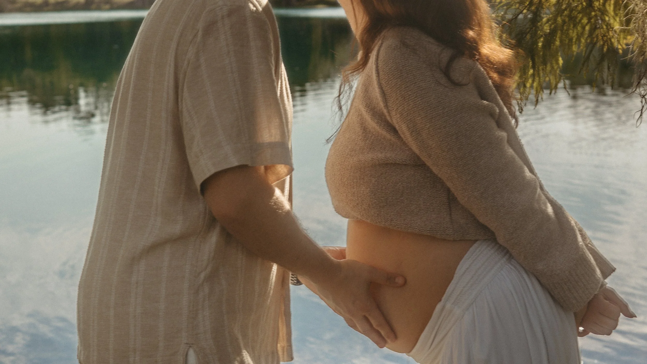 A couple standing close by a lake, with their faces out of the frame. The woman is in a beige sweater and white shorts, and the man is wearing a beige plaid shirt. The scene is bathed in warm, natural light.