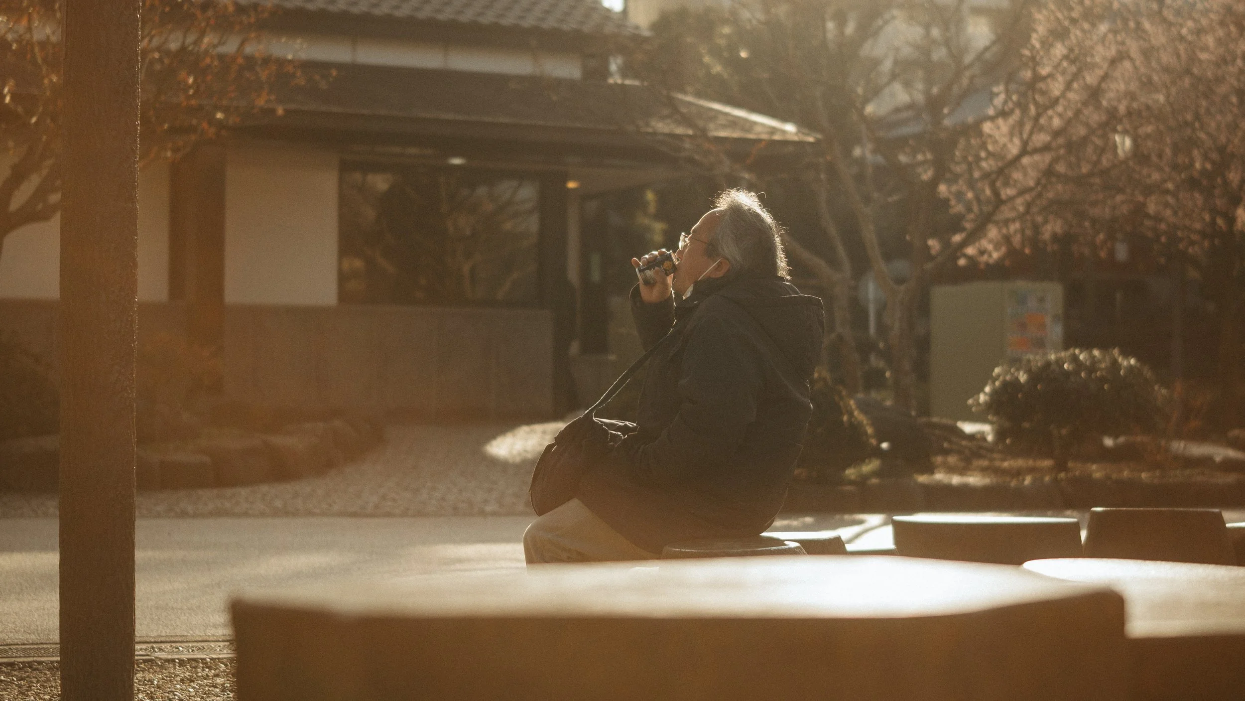 An elderly man with gray hair and glasses, wearing a face mask, sitting on a bench in a park during sunset, drinking from a can.