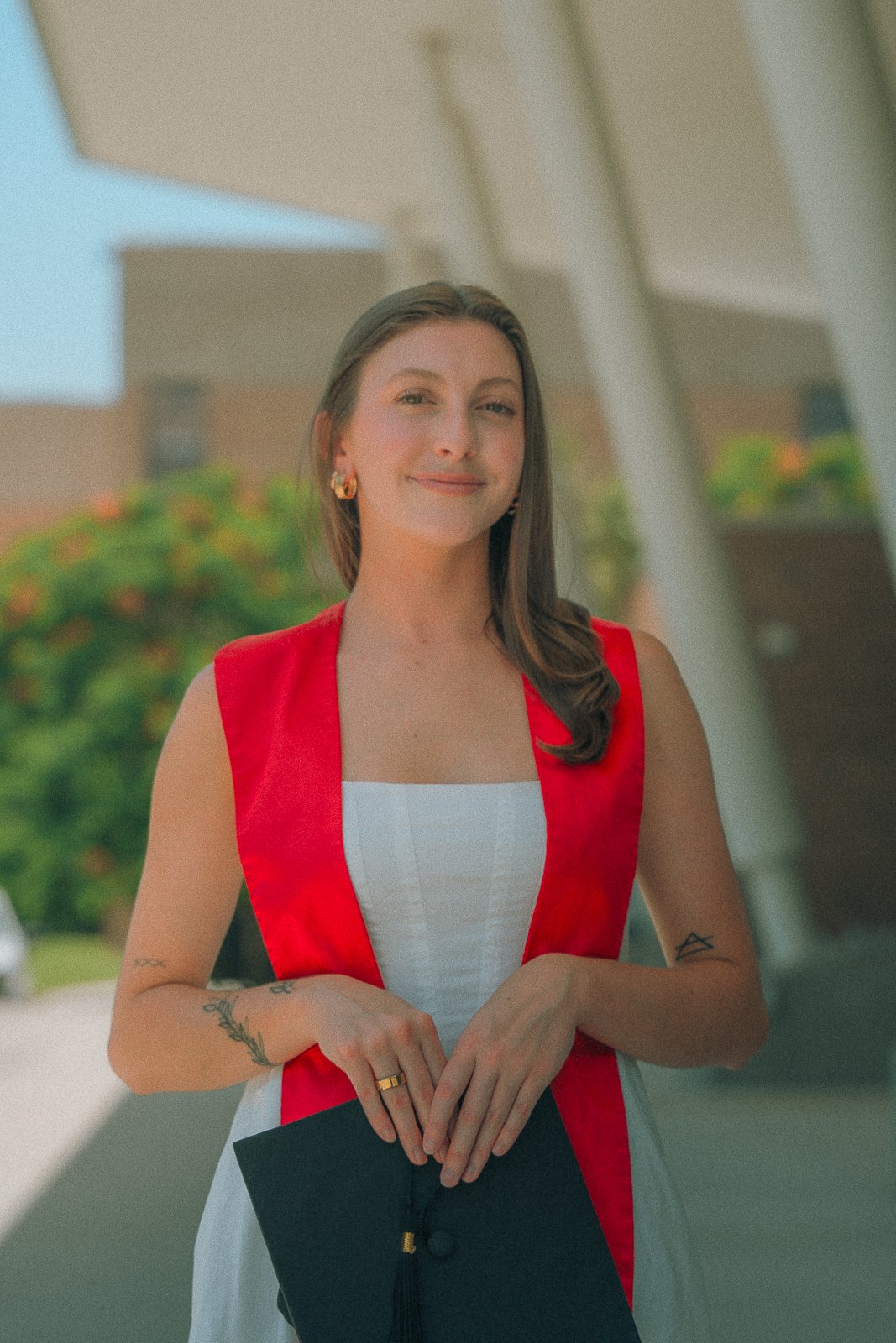 A young woman with long brown hair wearing a white dress with a red sash, holding a black folder, standing outdoors with a building and greenery in the background.