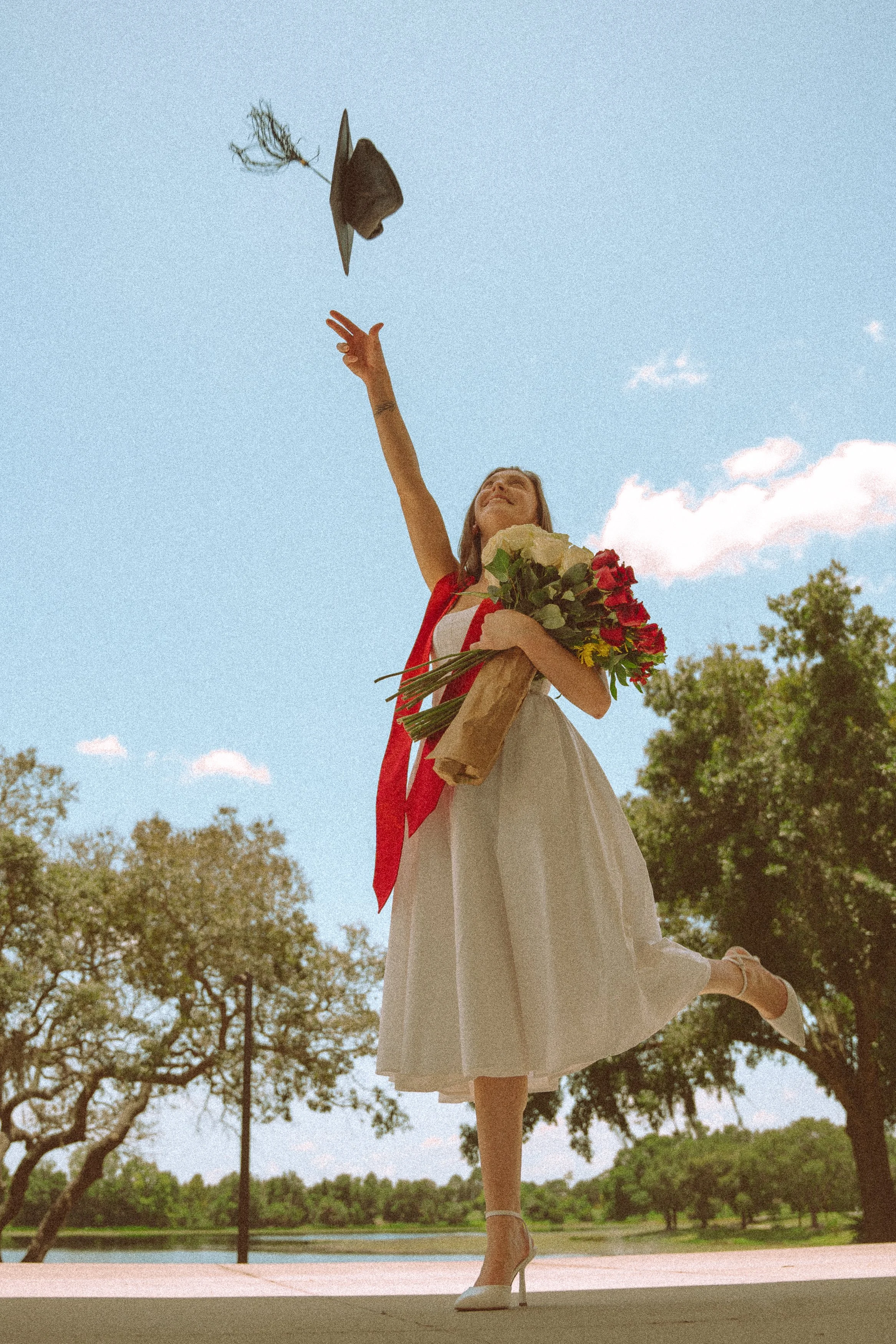 A woman in a white dress and high heels balancing on one foot outdoors, holding a large bouquet of red and white flowers with green foliage, with her other leg lifted behind her and her arm raised, tossing a hat into the air against a background of t