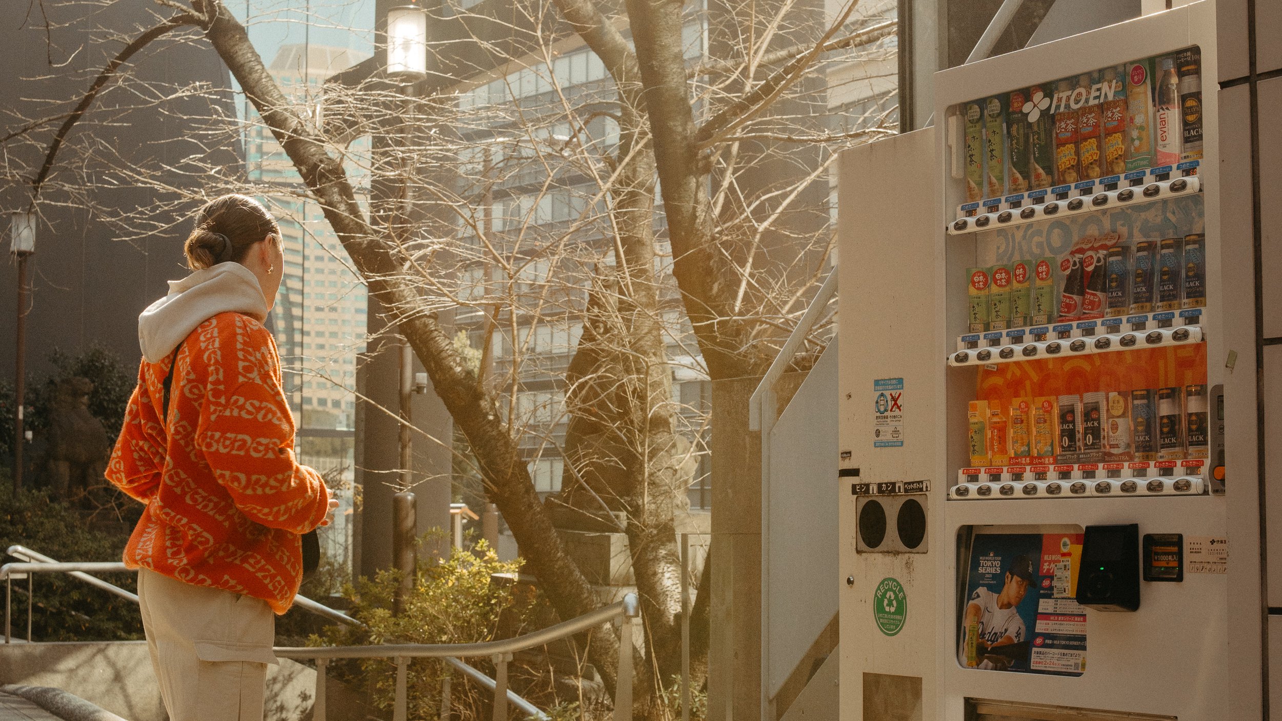 A person wearing a red hoodie with white writing and a white hoodie underneath, standing outside near a vending machine on a sunny day with leafless trees and tall buildings in the background.