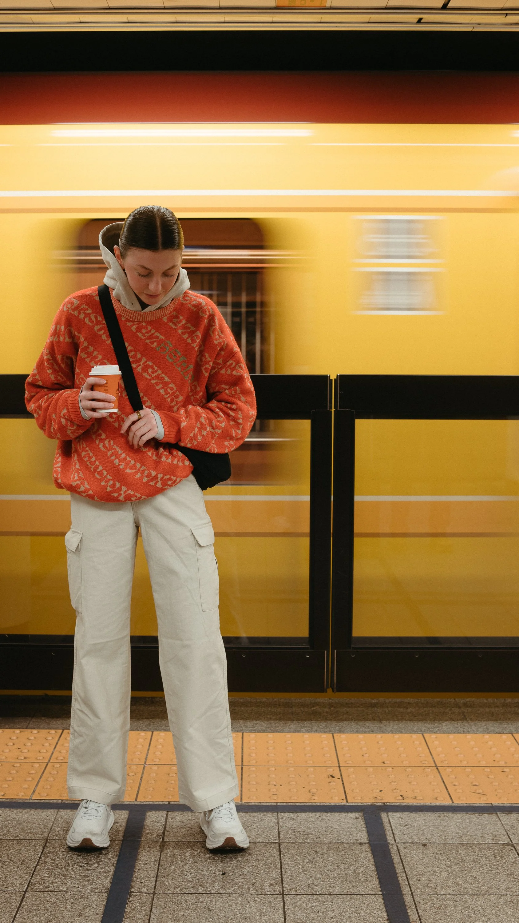 A young woman standing on a subway platform with a yellow subway train passing behind her, holding a coffee cup, dressed in a red patterned sweater and beige pants.