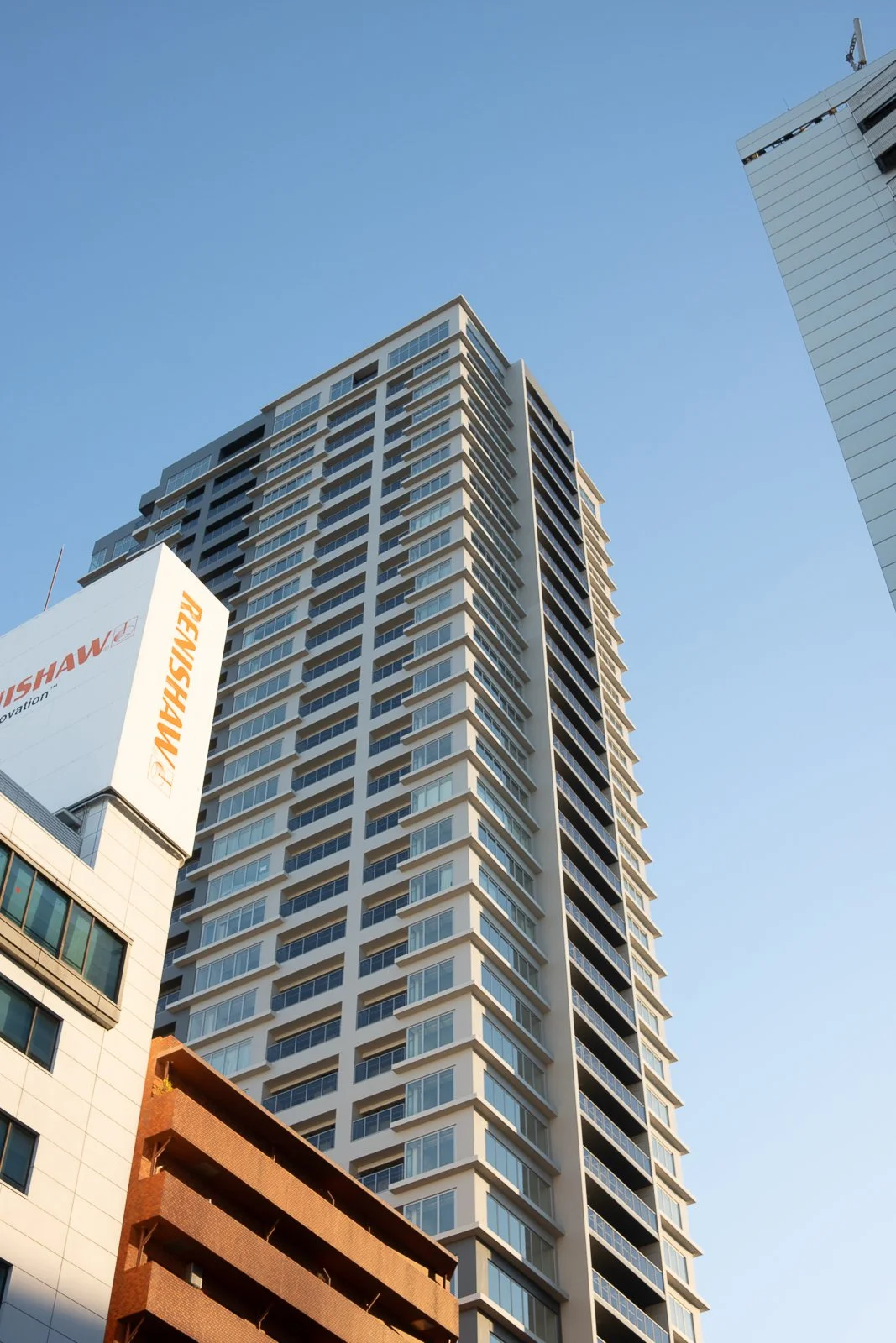 A tall modern high-rise building with numerous windows and balconies under a clear blue sky, with parts of other buildings visible nearby.