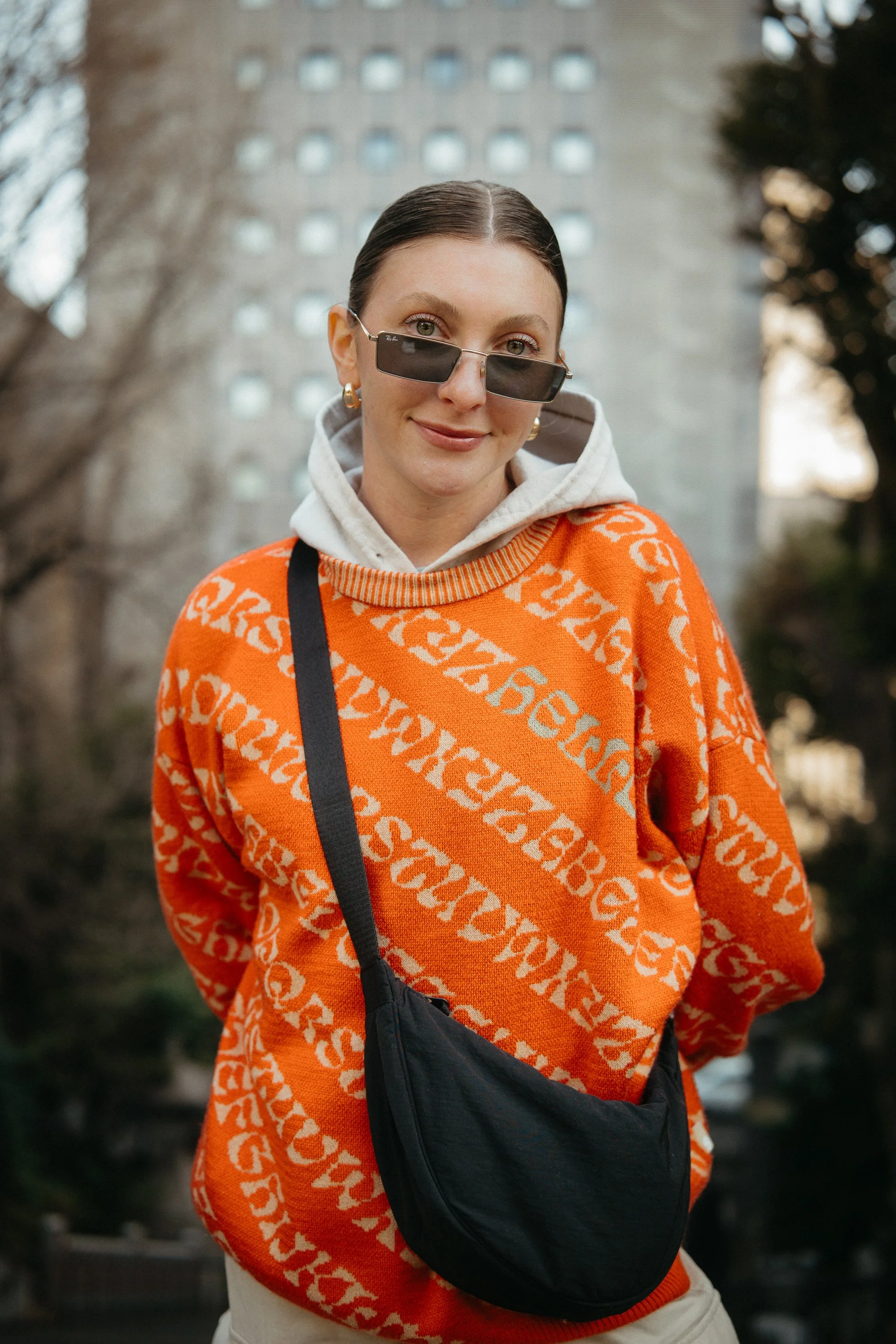 A woman with dark hair wearing sunglasses, a white hoodie, orange sweater with a white pattern, and gold hoop earrings, standing outdoors in front of trees and a tall building.