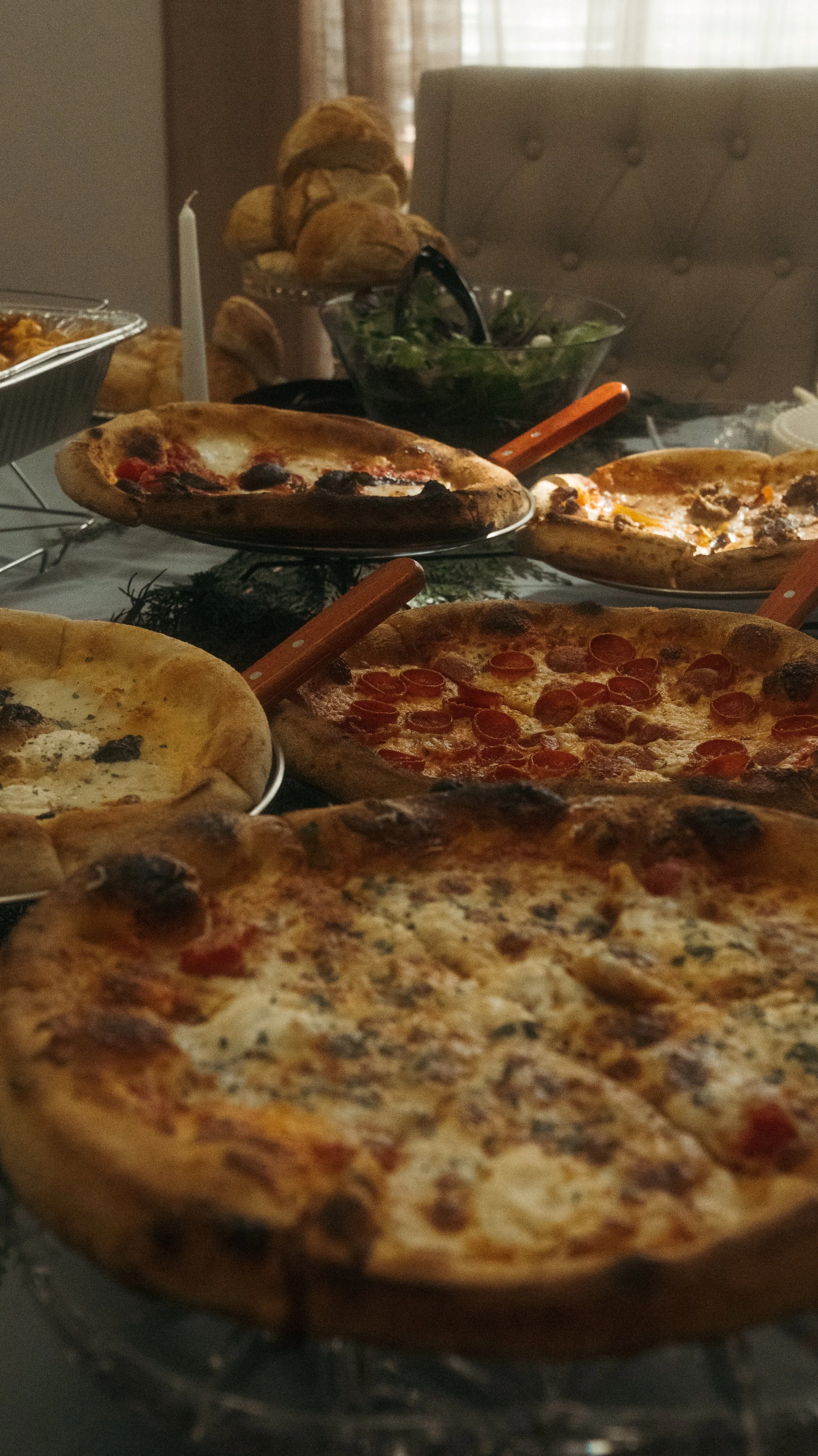 Several pizzas with various toppings are displayed on a table, with a salad and bread in the background.