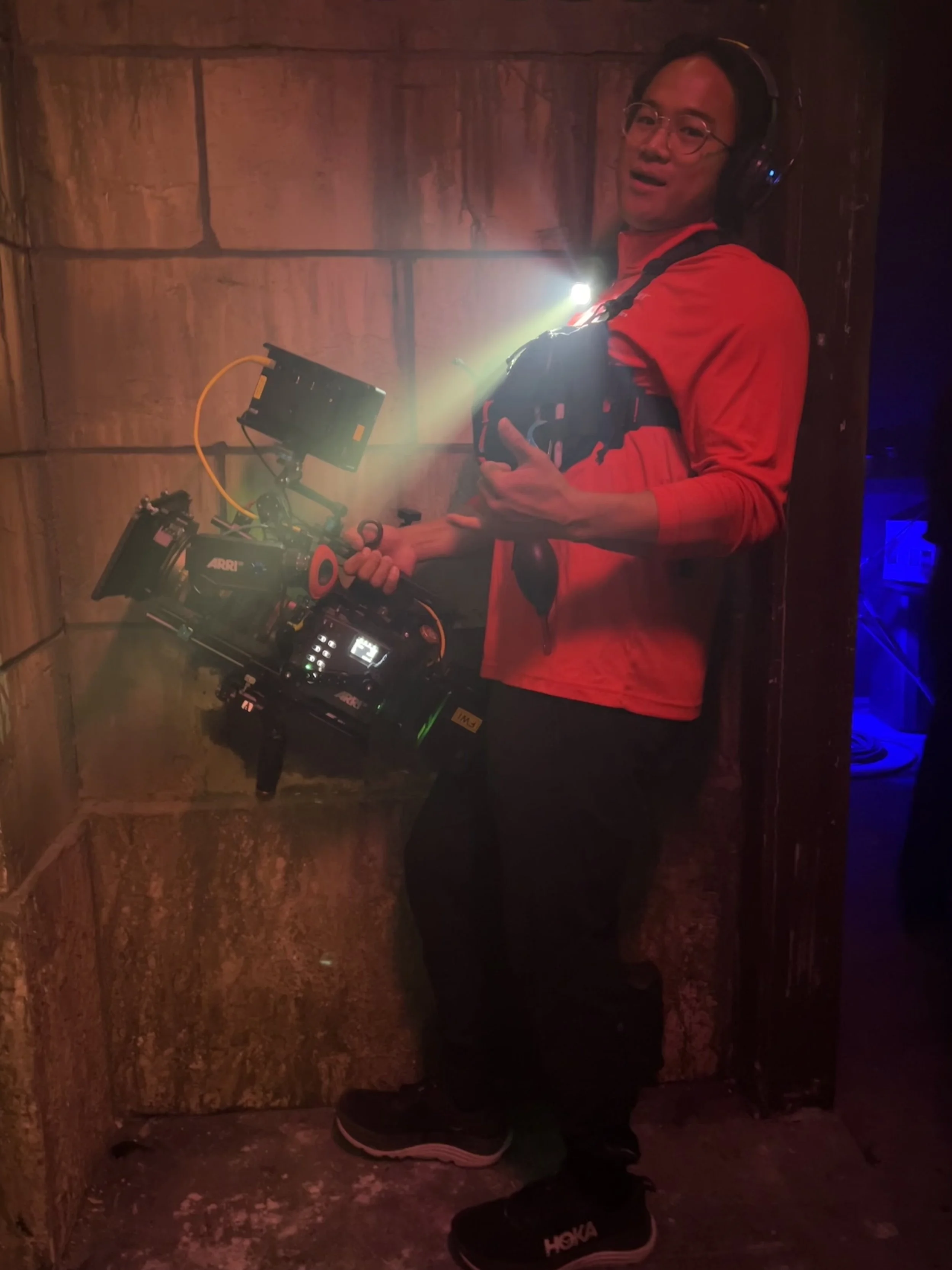 A person wearing glasses, a red jacket, and a black headlamp, holding a camera rig, standing against a wooden wall with their right hand pointing upwards.