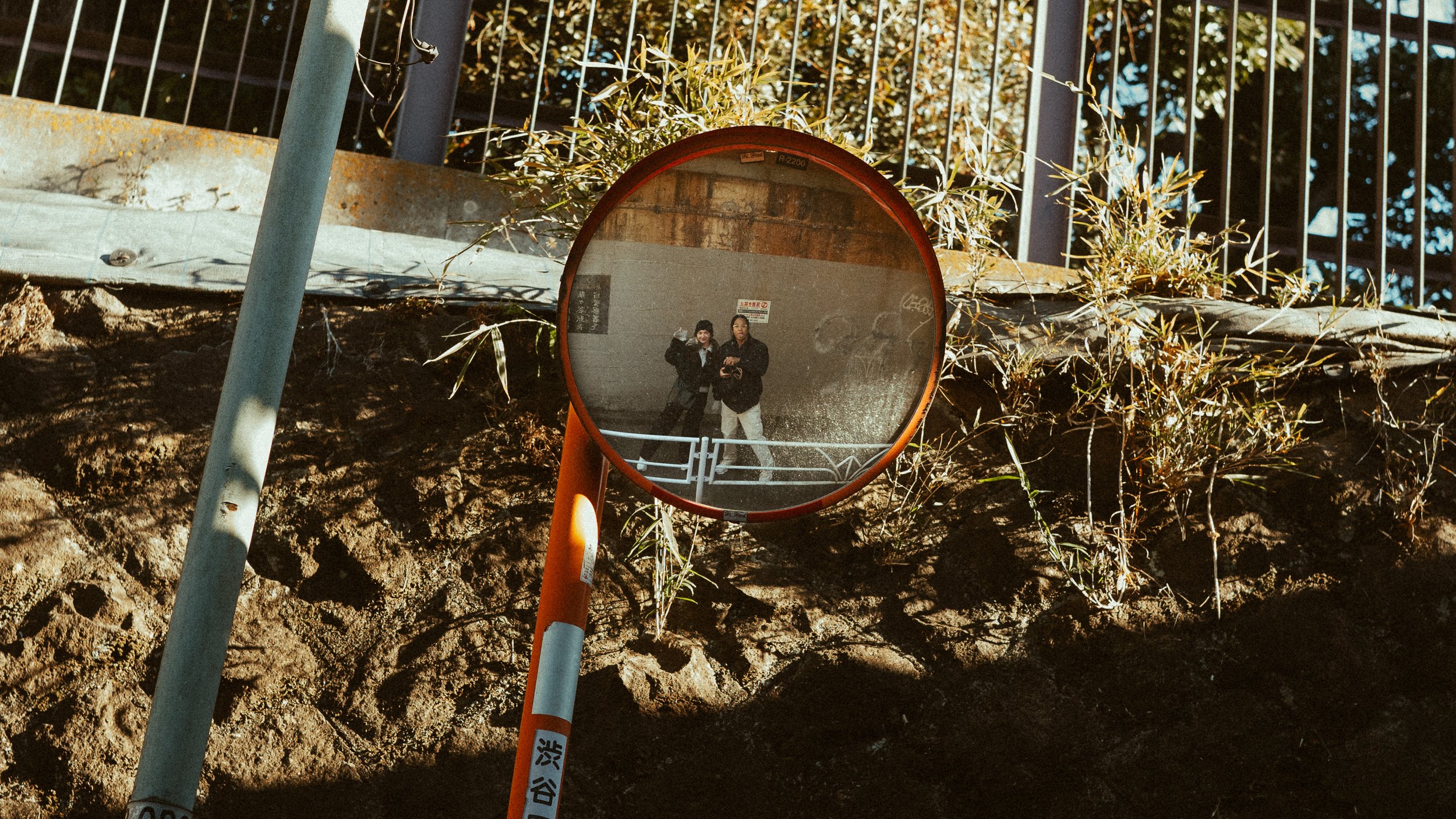Reflection of two people taking a photo in a round mirror mounted on a orange and white pole, outdoors with dirt ground and green plants, fence and blue sky in background.