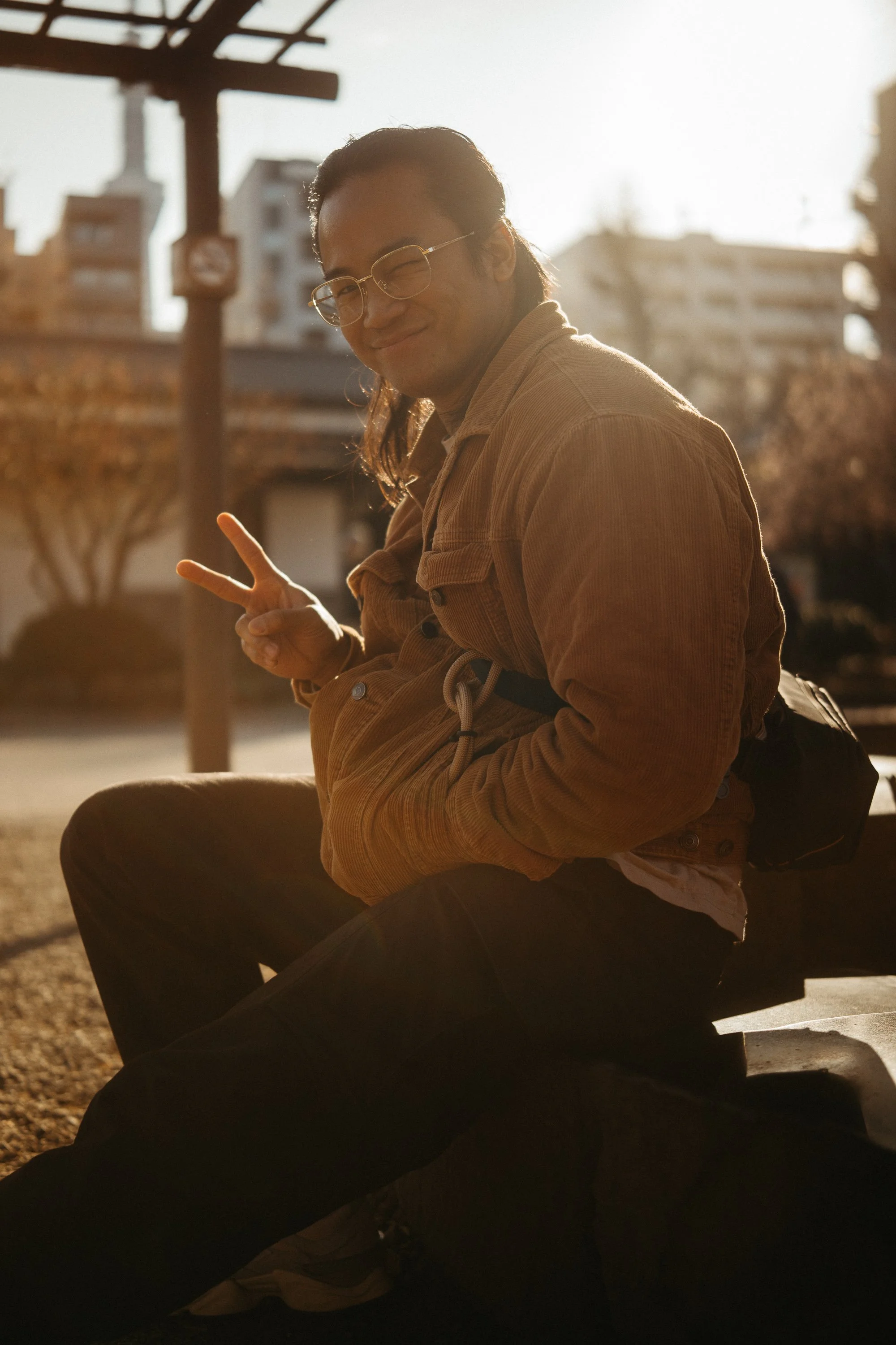 Person sitting outdoors in sunlight, smiling, making a peace sign with hand, wearing glasses and a brown jacket.
