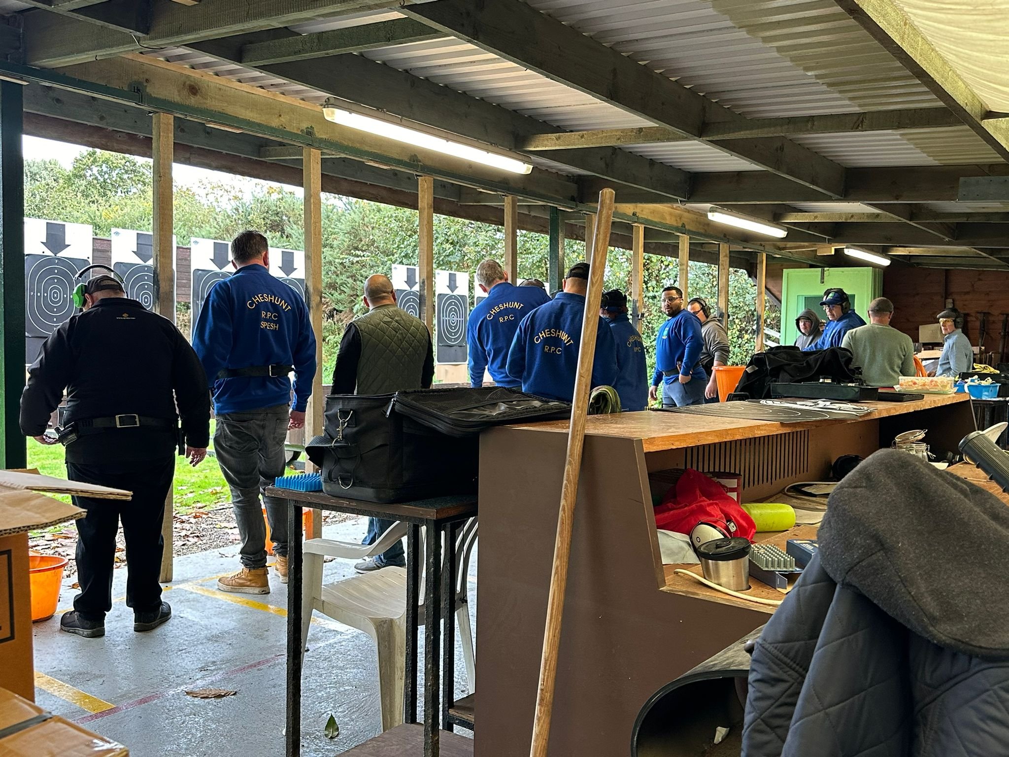 People participating in an outdoor shooting practice at a range, with targets set up at the back. The individuals are facing away from the camera, some wearing blue jackets with 'Cheshunt R.P.C.' on them. The setting appears to be a covered shooting pavilion with various equipment and supplies on the counter in the foreground.
