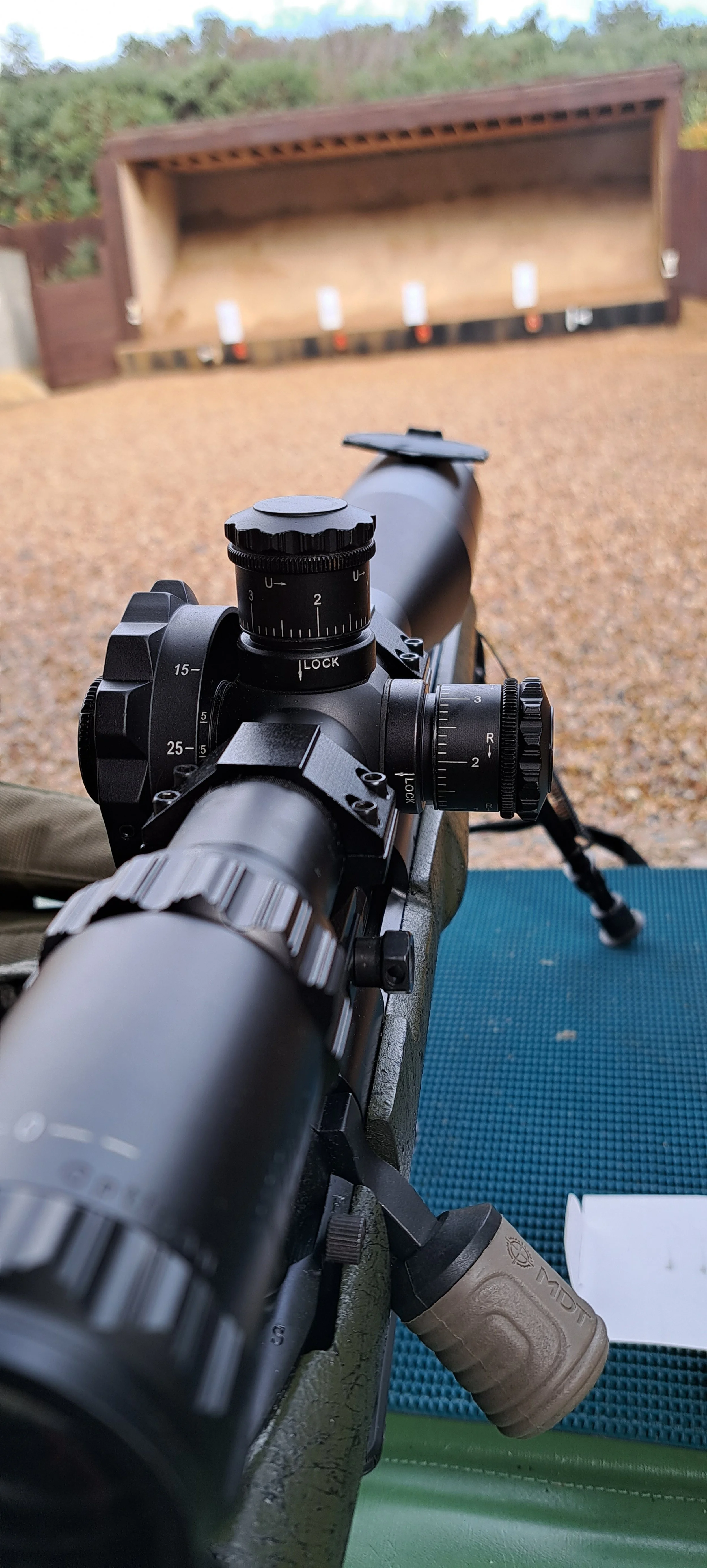 A scoped rifle aimed at shooting targets on a range with a wooden backstop, outdoors during daytime.