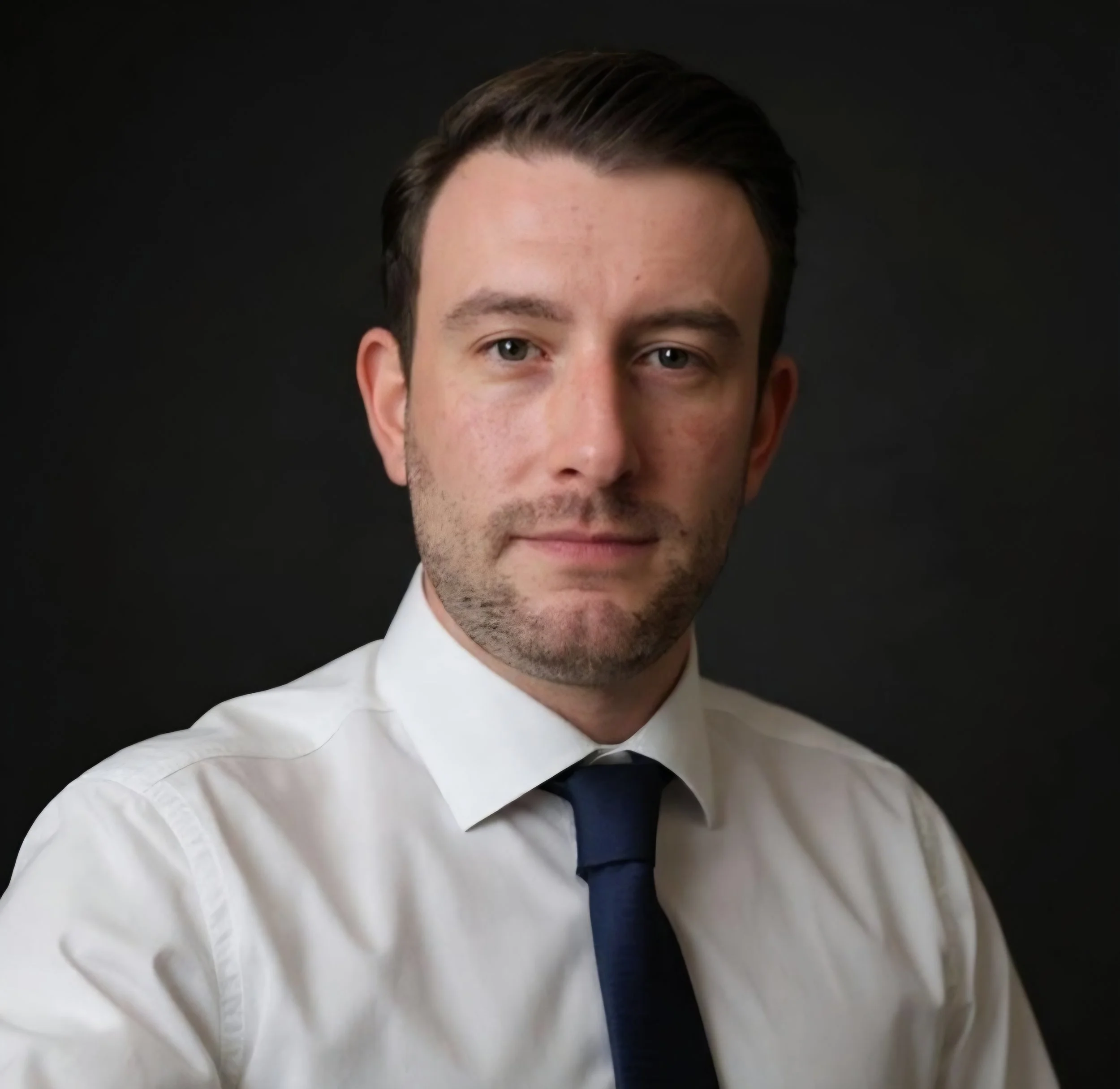 A portrait of a young man with short dark hair, wearing a white dress shirt and a dark blue tie, against a black background.