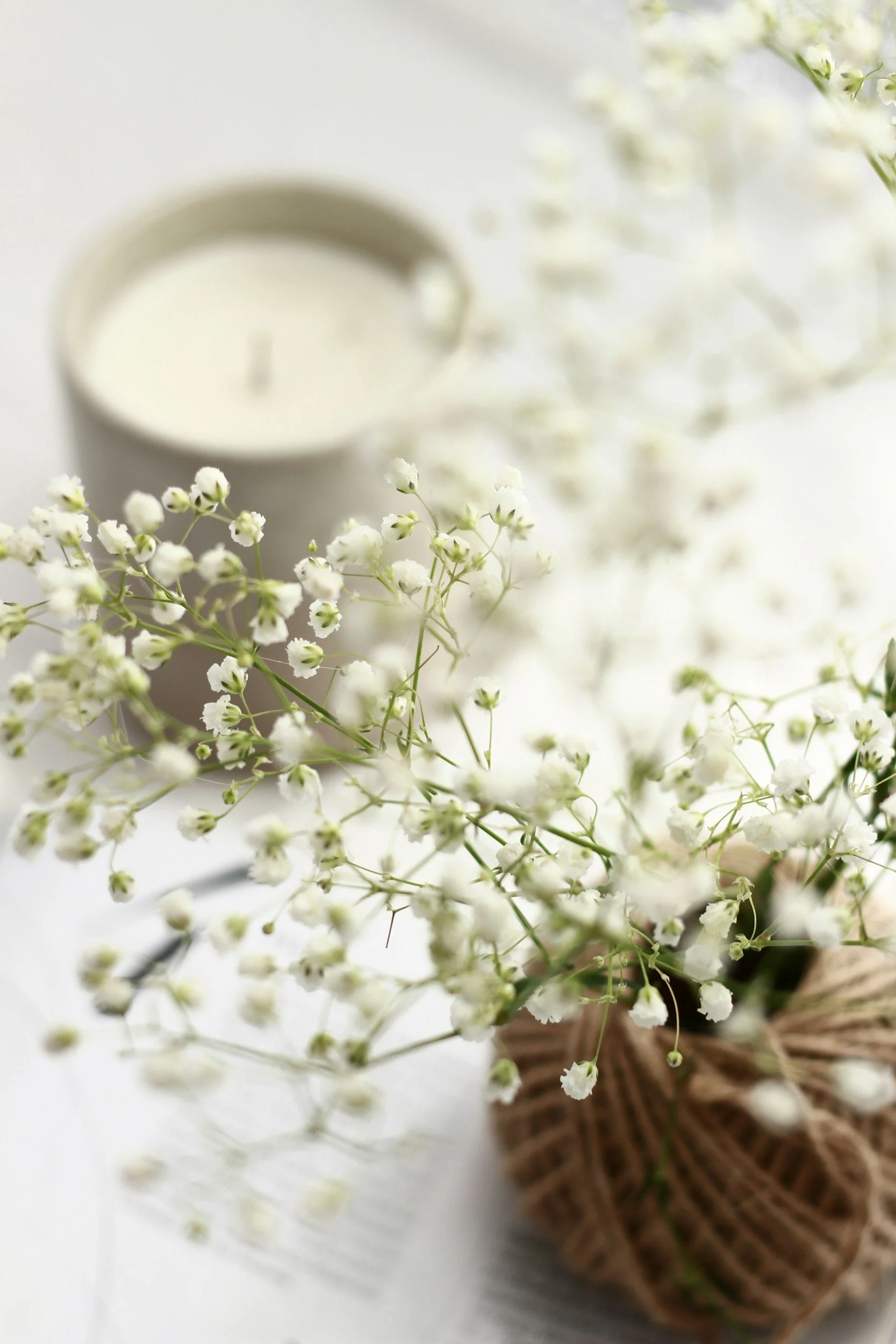 A bouquet of small white flowers wrapped in brown paper, a white candle, and a book on a white surface.