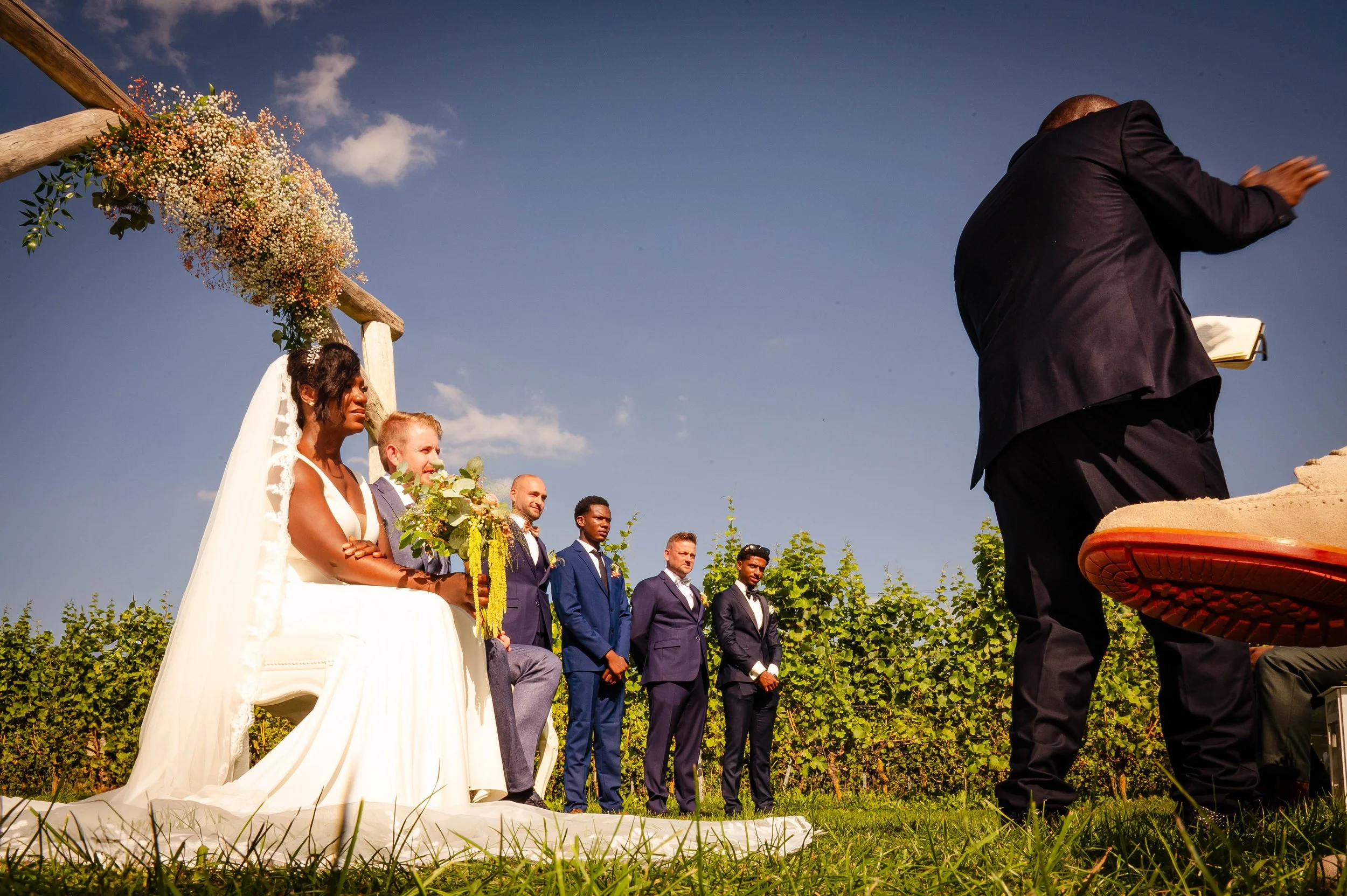 Een bruidsmeisje en een bruidegom zitten onder een bloemenboog tijdens een buitenbruiloft, terwijl de ceremoniemeester of dominee voor hen staat met een boek in de hand.