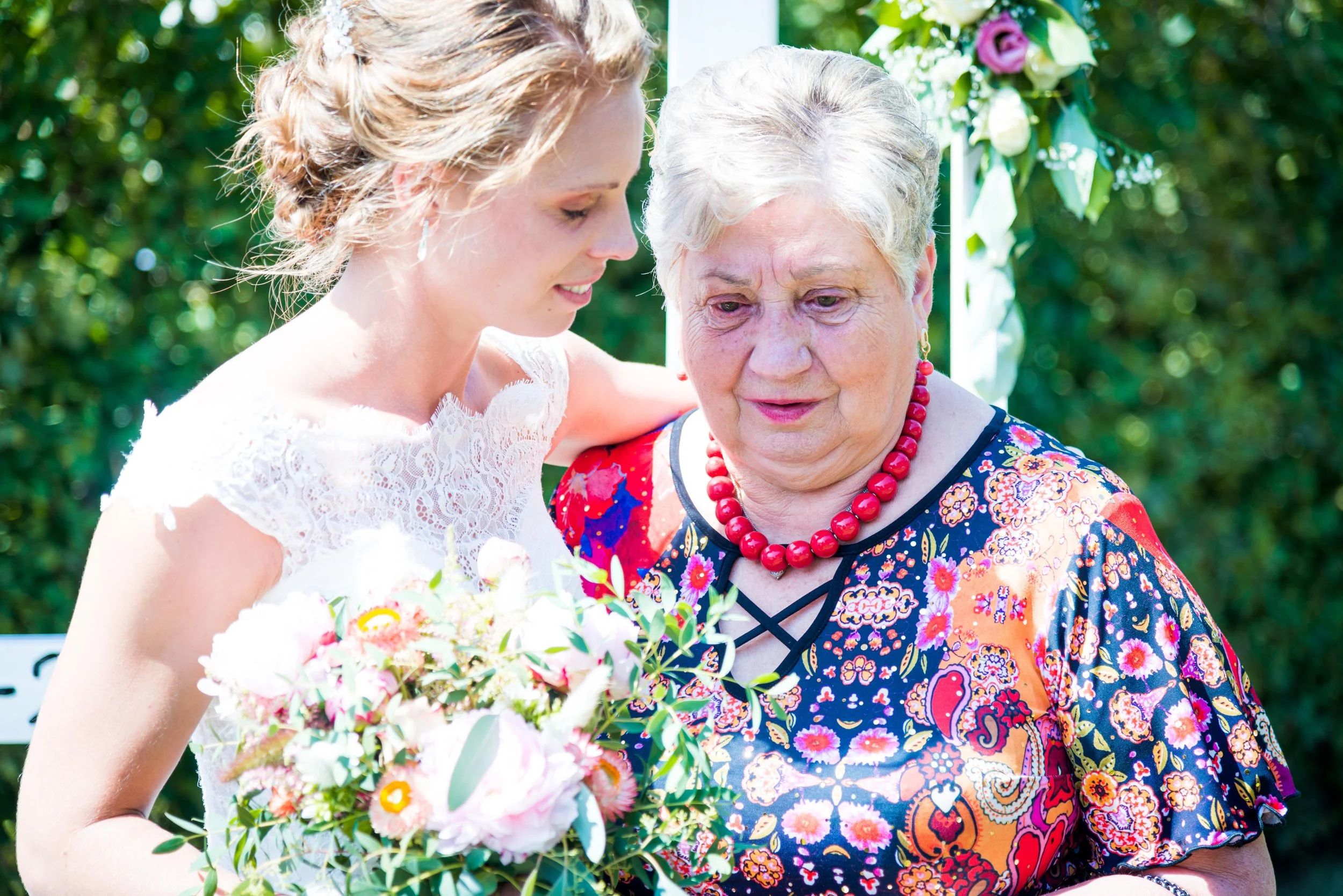 Een jonge vrouw in een witte jurk en een oudere vrouw met een kleurrijke jurk en een rode ketting, knuffelen en kijken naar een bloemenboeket tijdens een buitengebeurtenis.