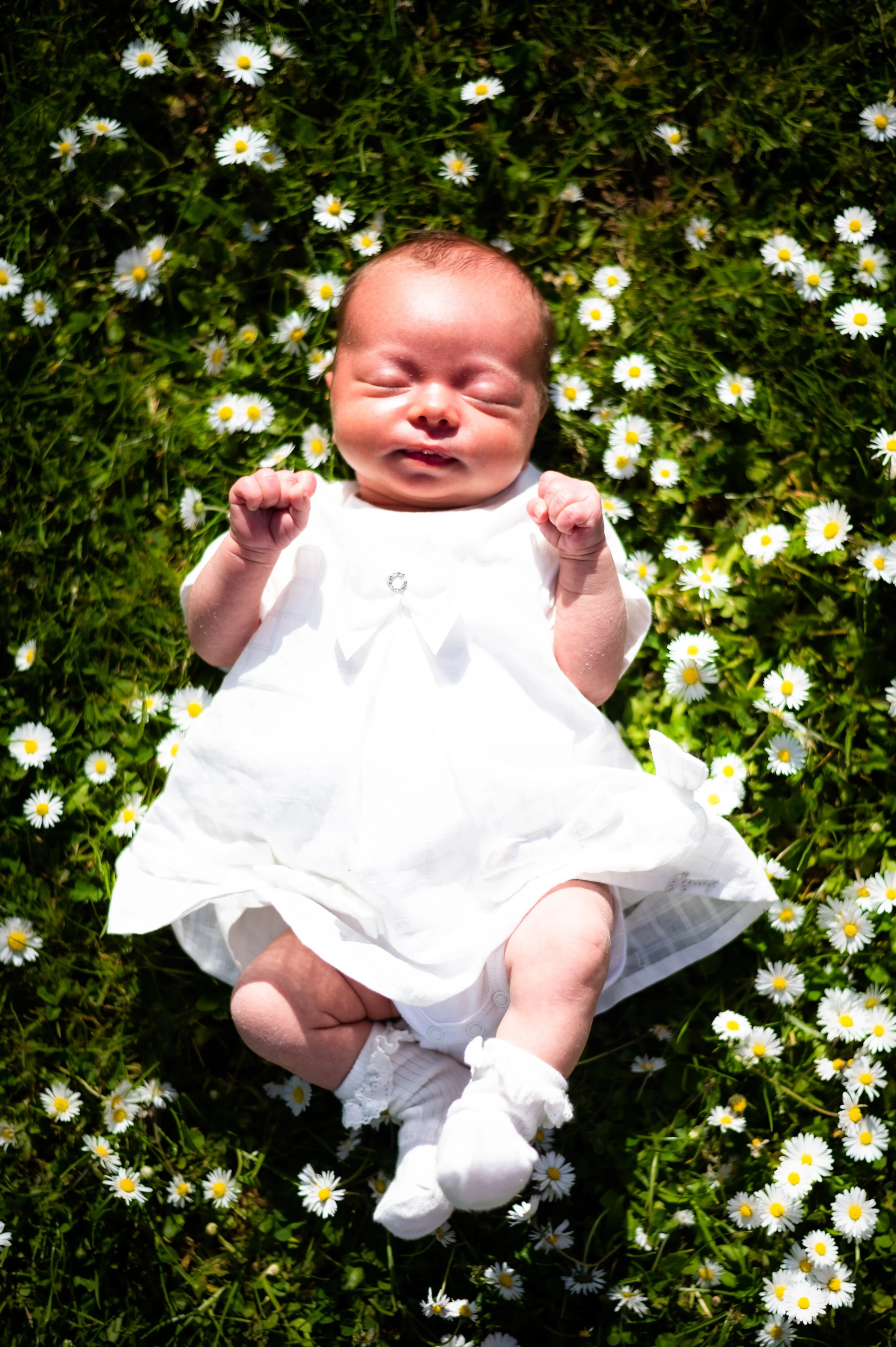 Baby in witte jurk ligt op grasveld met madeliefjes.