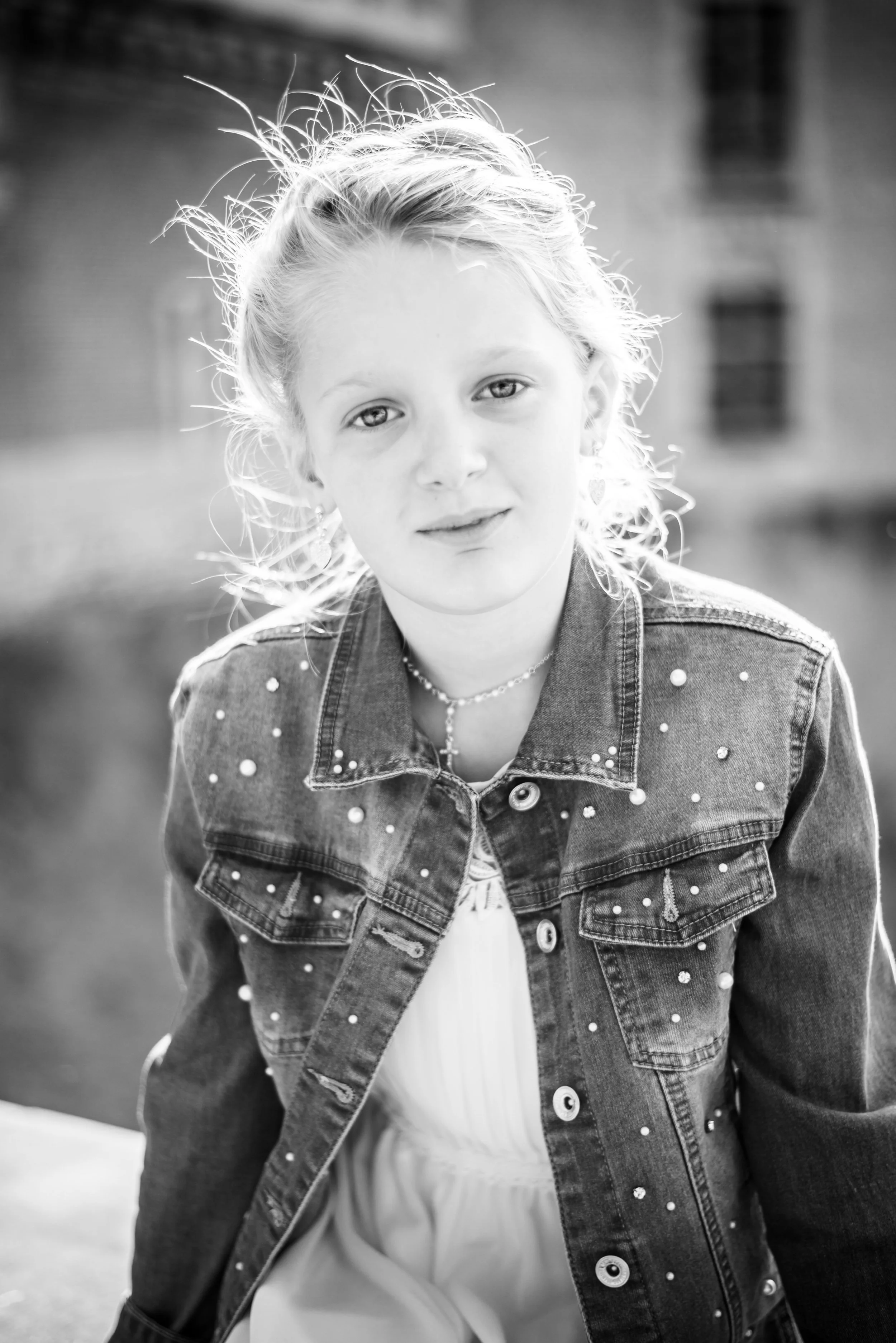 Zwart-wit foto van een jonge meisje met warrig haar, gekleed in een denim jas met parels, aangeraakt door zacht backlight.