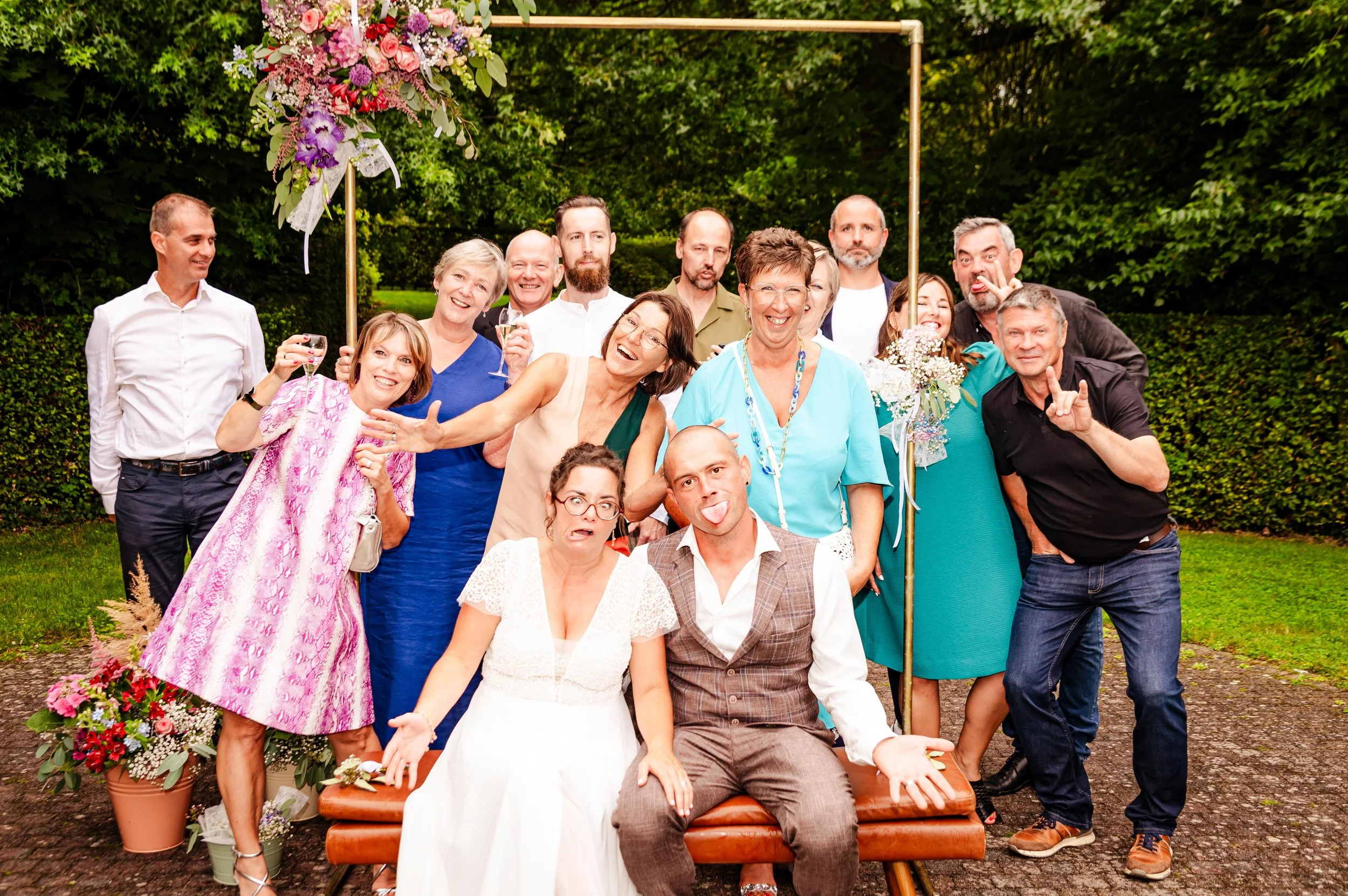 Groep mensen poseren bij een tuinbruiloft, met een bruid en bruidegom zittend vooraan, omringd door familie en vrienden, met bloemen en groene achtergrond.