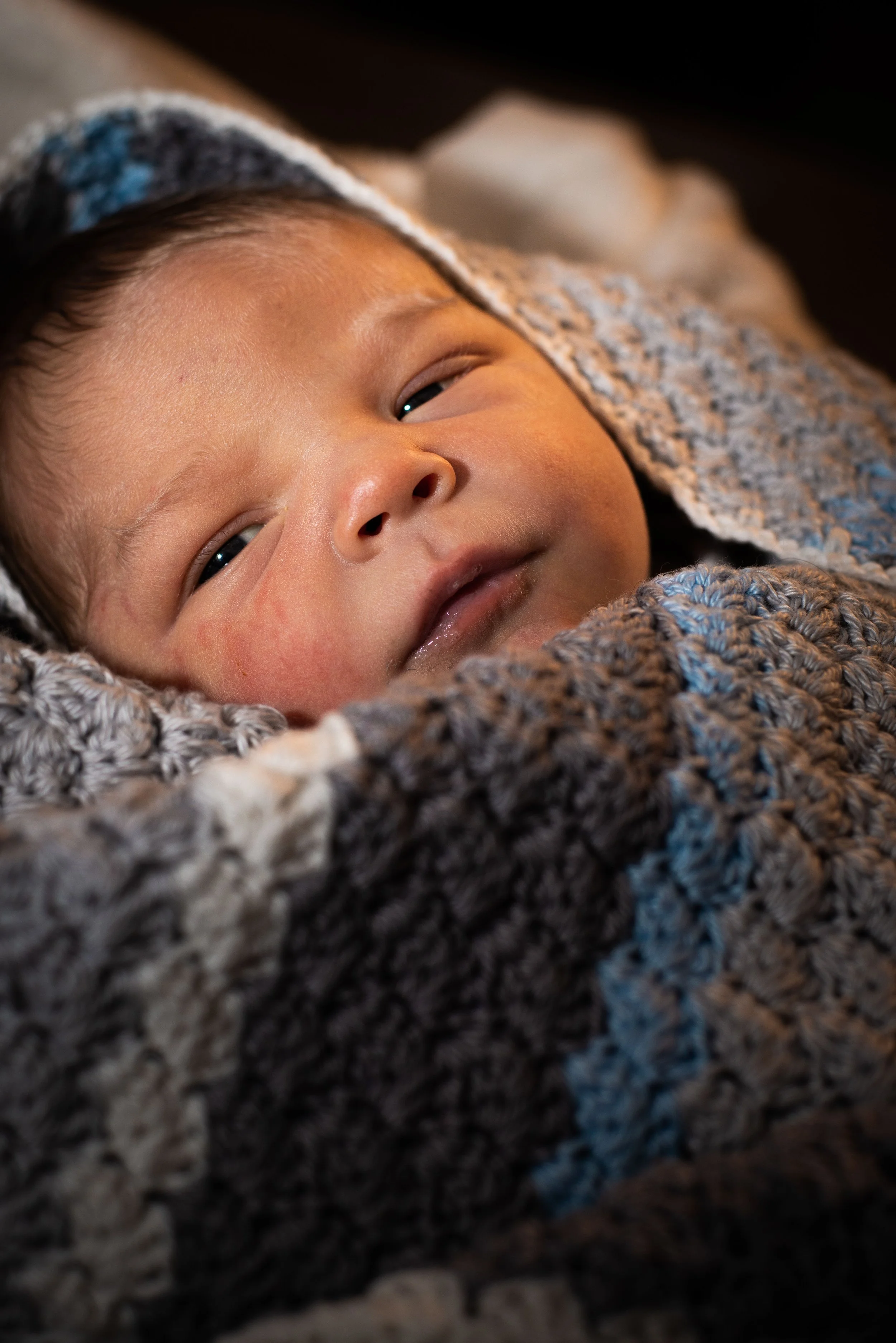 Een baby ligt onder een gebreide deken en kijkt nieuwsgierig naar de camera.