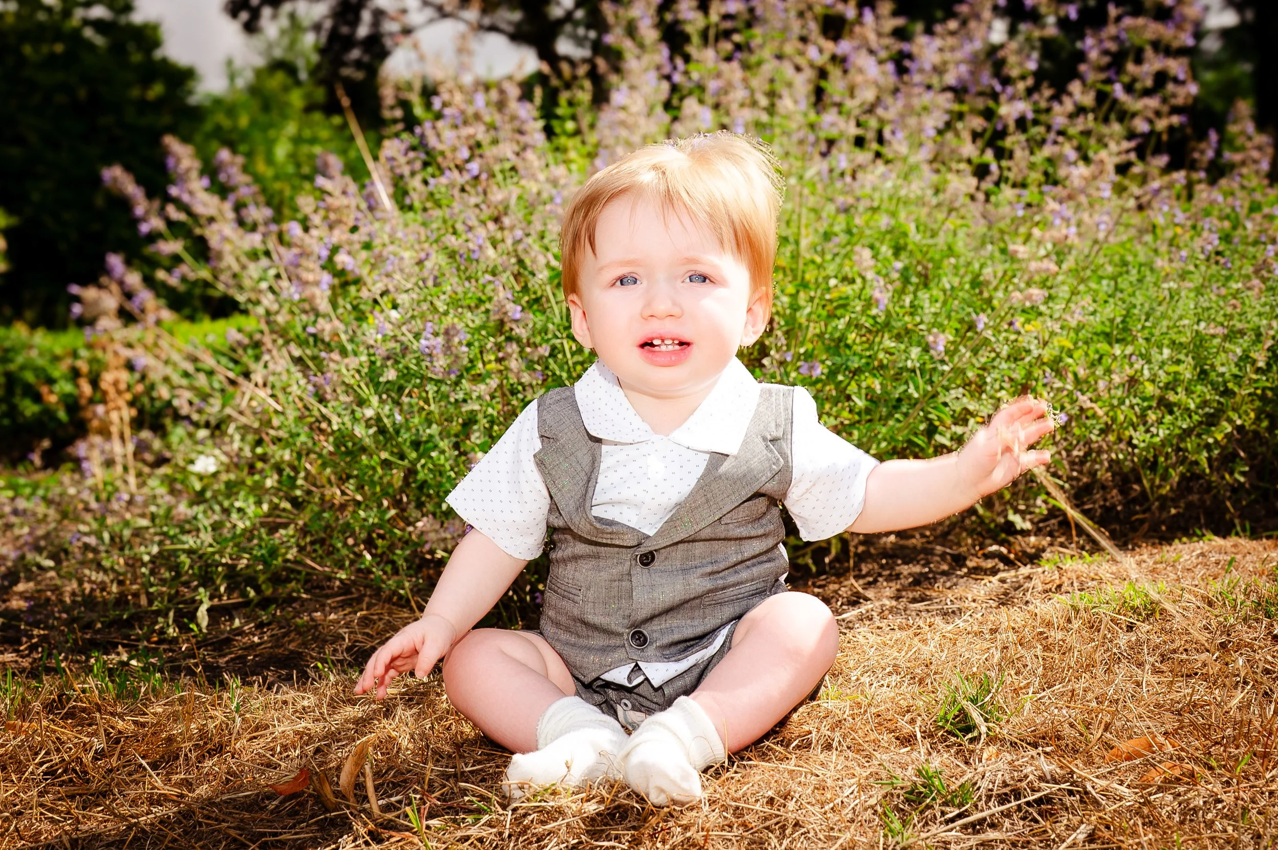 Kleine jongen in nette kleding zittend op de grond, omgeven door bloemen en groen, in een buitenomgeving.