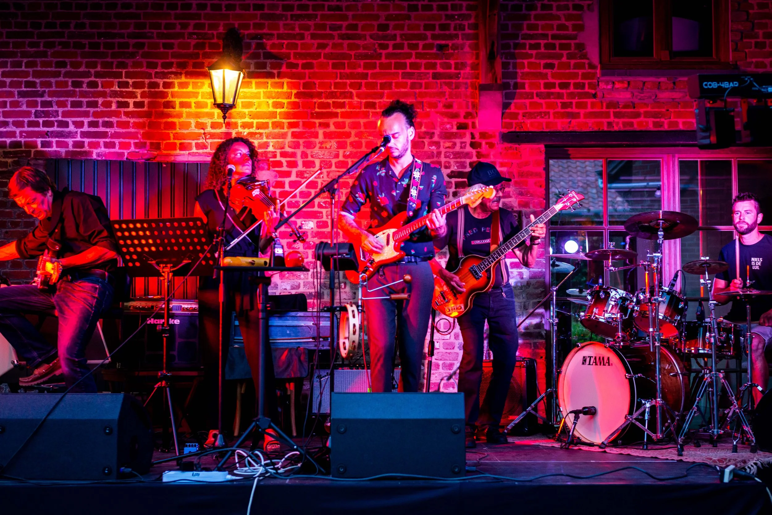 Een band met meerdere muzikanten die live op een podium optreden, onder kleurrijk stageverlichting, met een bakstenen muur op de achtergrond.