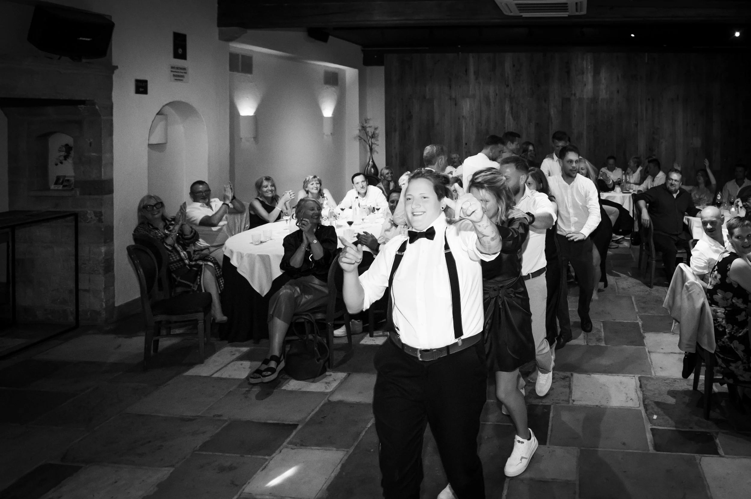 Grup mensen danst en lacht in een feestzaal, met een publiek die applaudisseert en toekijkt.