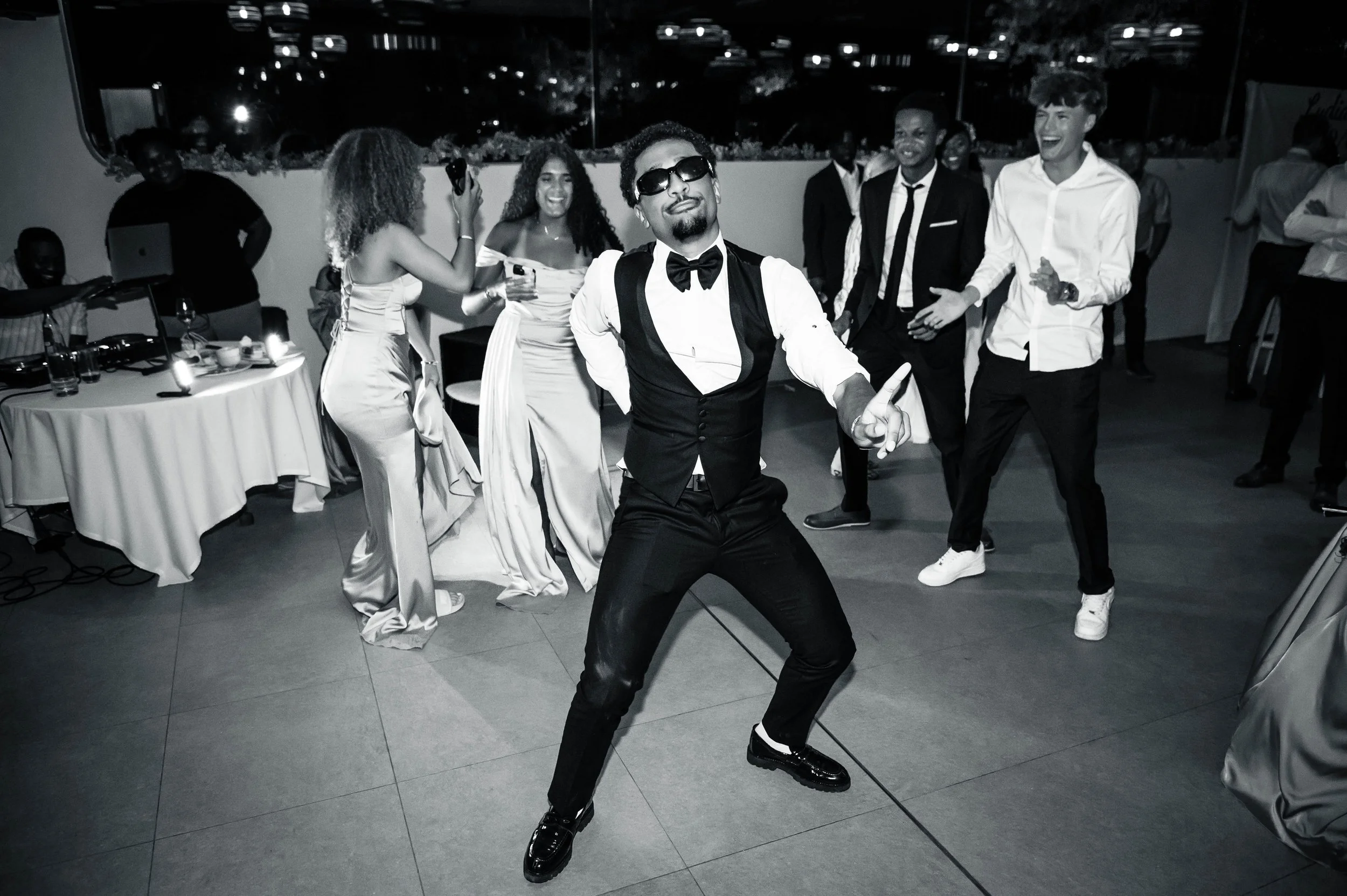 Mensen dansen en lachen op feest met stijlvolle kleding, een man in een pak met zonnebril in het centrum.