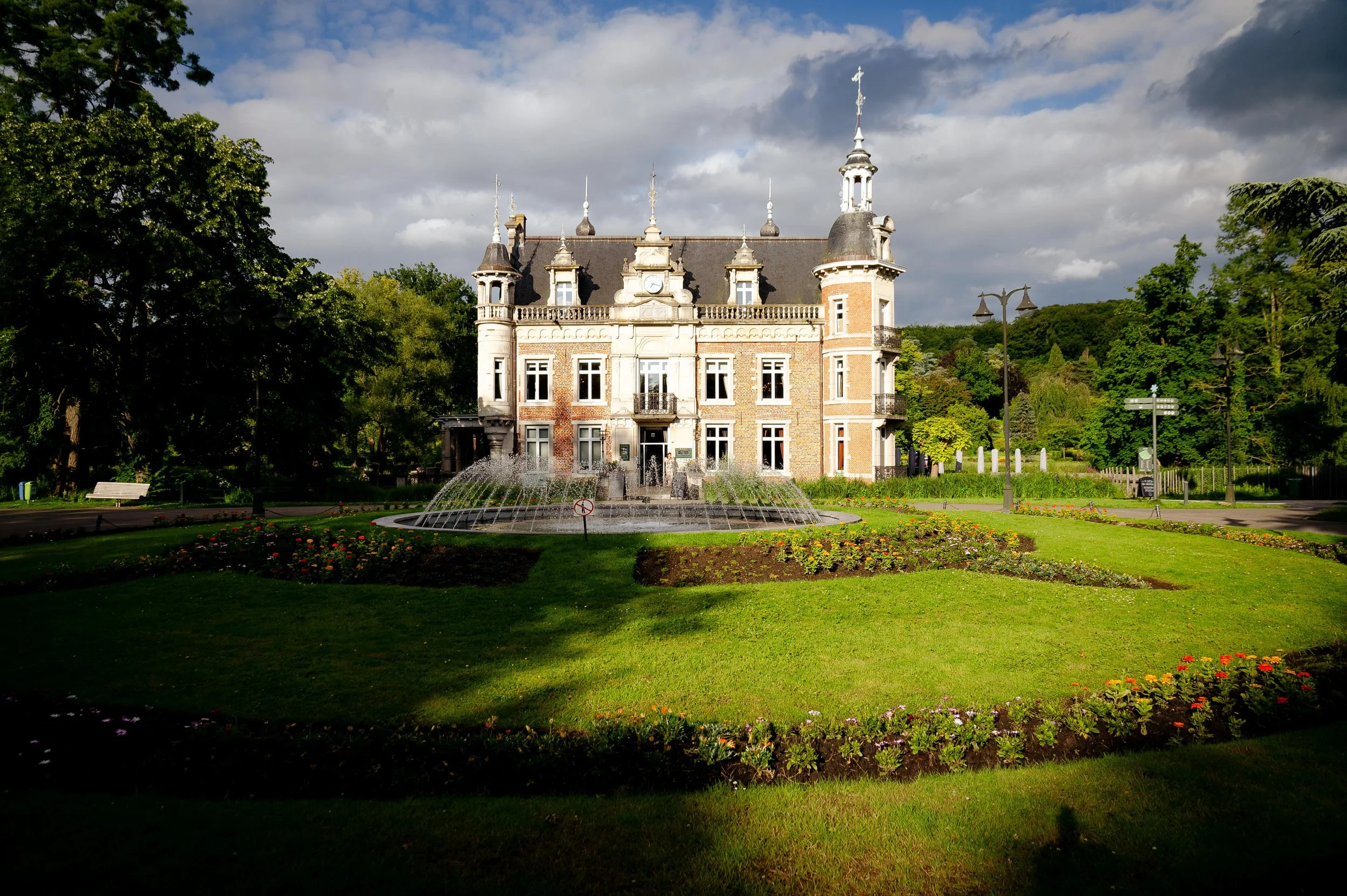 Een historisch kasteel met een waterfontein op de voorgrond, omgeven door een tuin met bloemen en bomen, onder een bewolkte hemel.