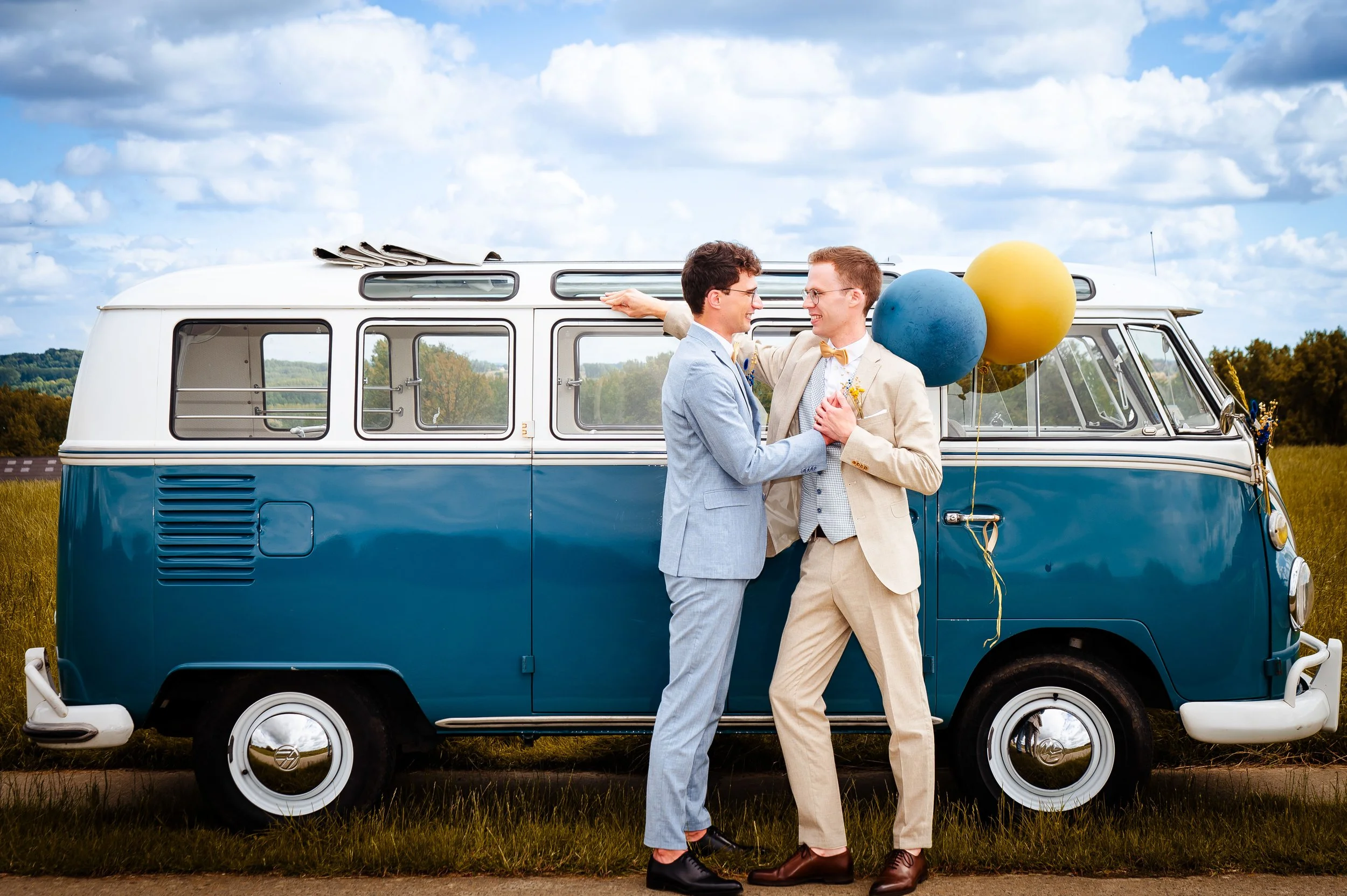 Twee mannen die elkaar liefkozen naast een blauwe en witte vintage bus met ballonnen op het platteland.