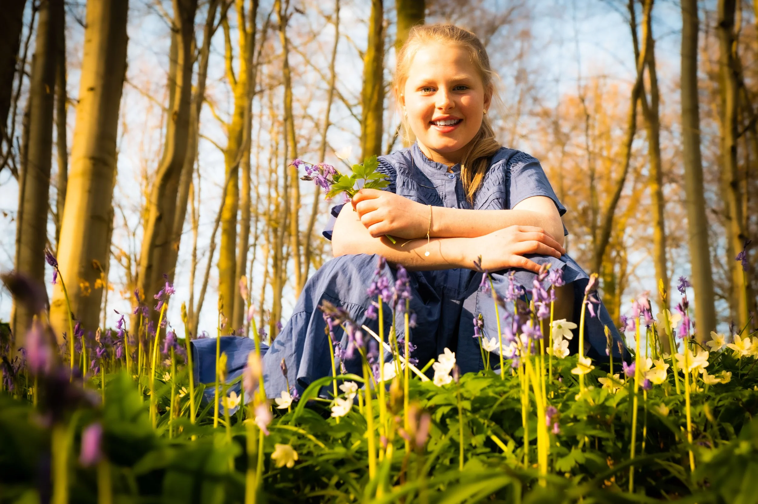 Lachende jonge vrouw zittend op de grond in een bos, omringd door paarse en witte bloemen, met gevallen bladeren aan de bomen op de achtergrond.