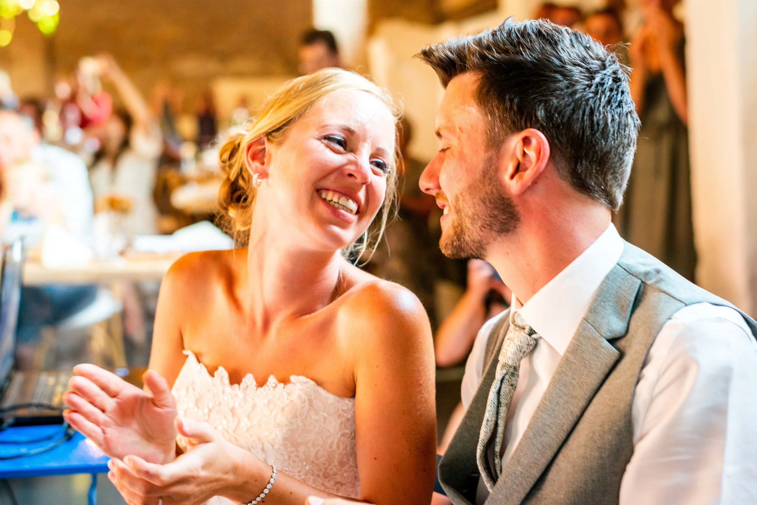 Een gelukkige bruid en bruidegom die elkaar aankijken en lachen tijdens hun huwelijk, omringd door gasten in een feestzaal.