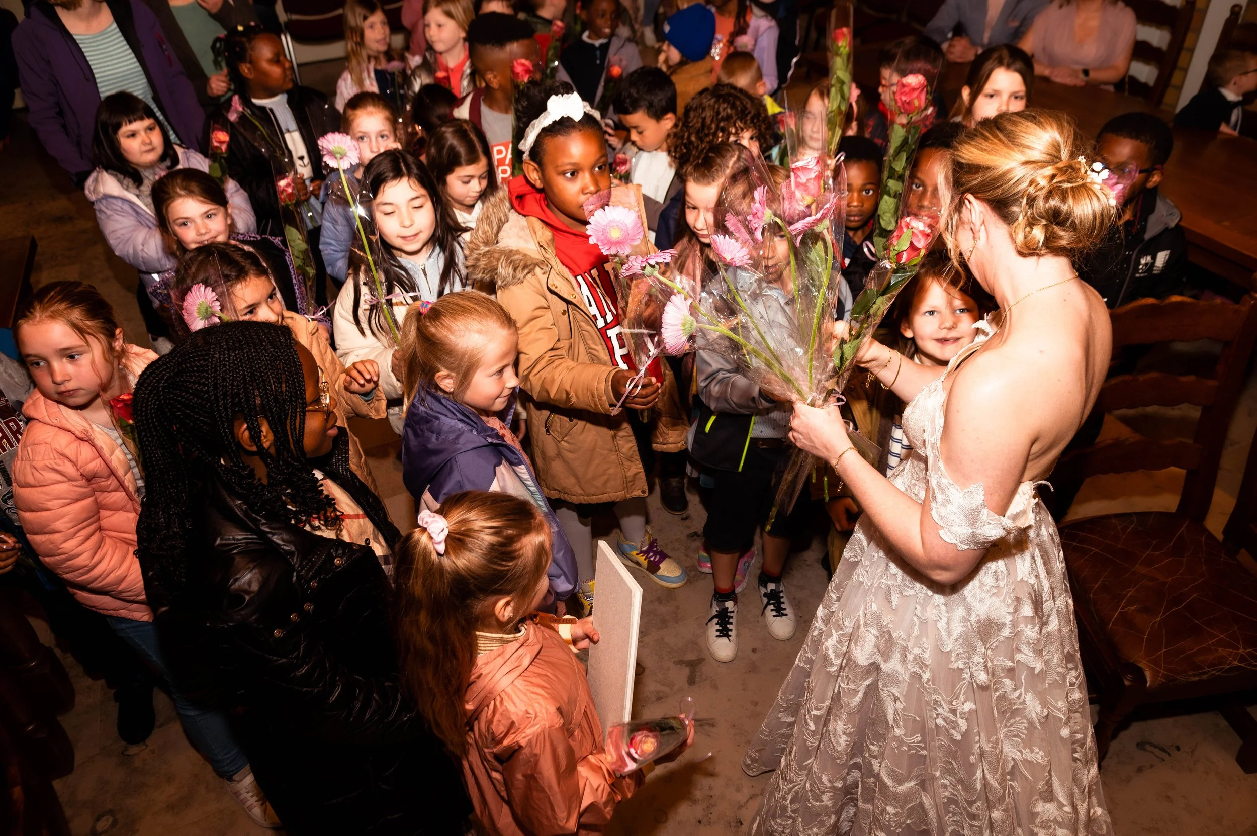 Een vrouw in een witte jurk geeft bloemen aan een groep kinderen die in een rij staan. De kinderen wachten in een zaal en kijken naar de vrouw, sommigen glimlachen.