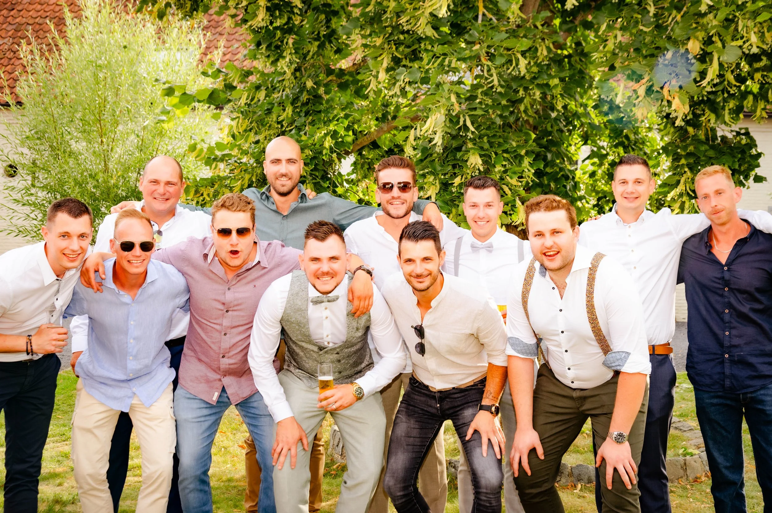 Een groep van 13 mannen die samen lachen en poseren voor een foto buiten, onder een boom, tijdens een feest of bijeenkomst, in zomerse kleding.