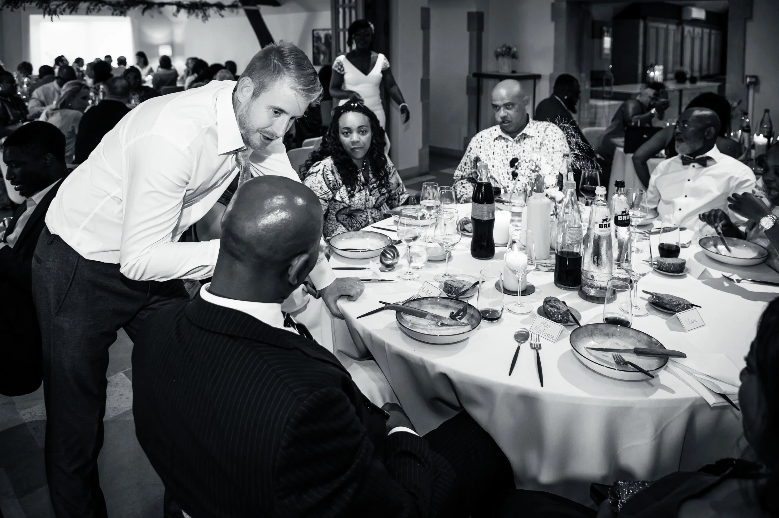 Mensen zitten aan een gedekte feesttafel in een restaurant of banquetzaal, met verschillende drankjes, borden en bestek. Eén man staat en wijst naar een ander die aan tafel zit, terwijl anderen kijken of praten.