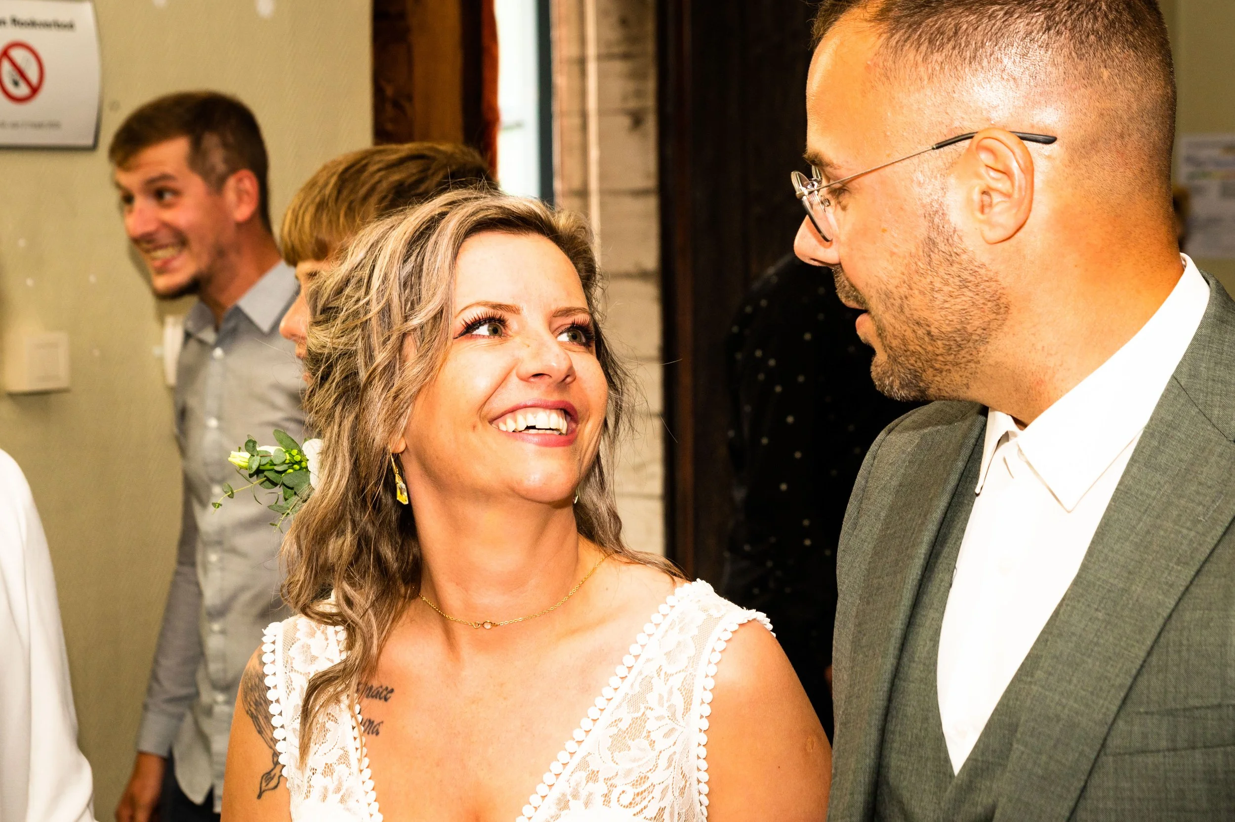 Een vrouw en een man die naar elkaar kijken en glimlachen tijdens een feest of gebeurtenis, met andere mensen op de achtergrond.
