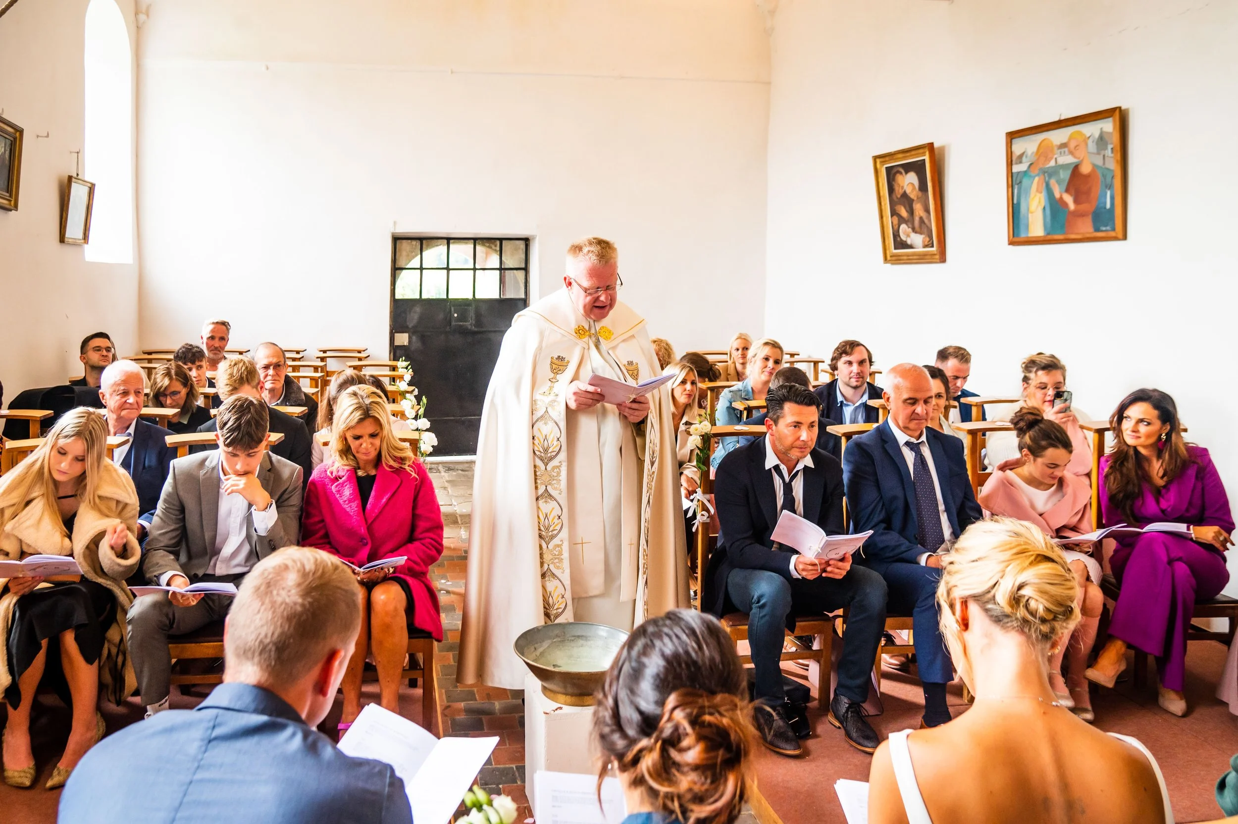 Een plechtige huwelijksceremonie in een kerk, met een priester die voor de bruidegom en bruid spreekt. De gasten zitten in banken, lezen of kijken. De kerk heeft witgekalkte muren, raamdecoraties en schilderijen aan de muur.