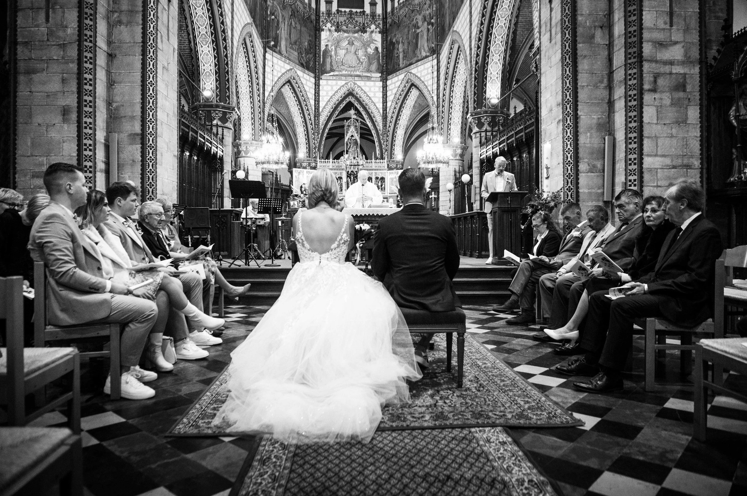 Een zwart-wit foto van een huwelijksmis in een gotische kerk. De bruid en bruidegom zitten op een bank in het midden, omringd door familie en vrienden die de ceremonie bijwonen.