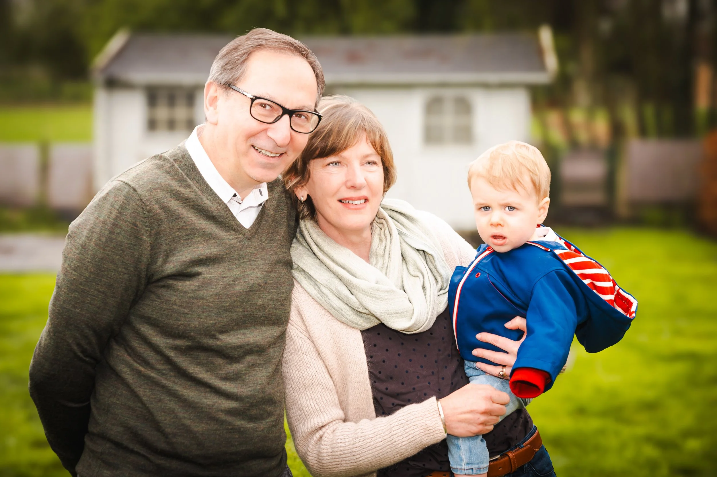 Familie van drie mensen buiten in een tuin, met een huis en groen landschap op de achtergrond. De man glimlacht, de vrouw houdt een jonge jongen in haar armen.