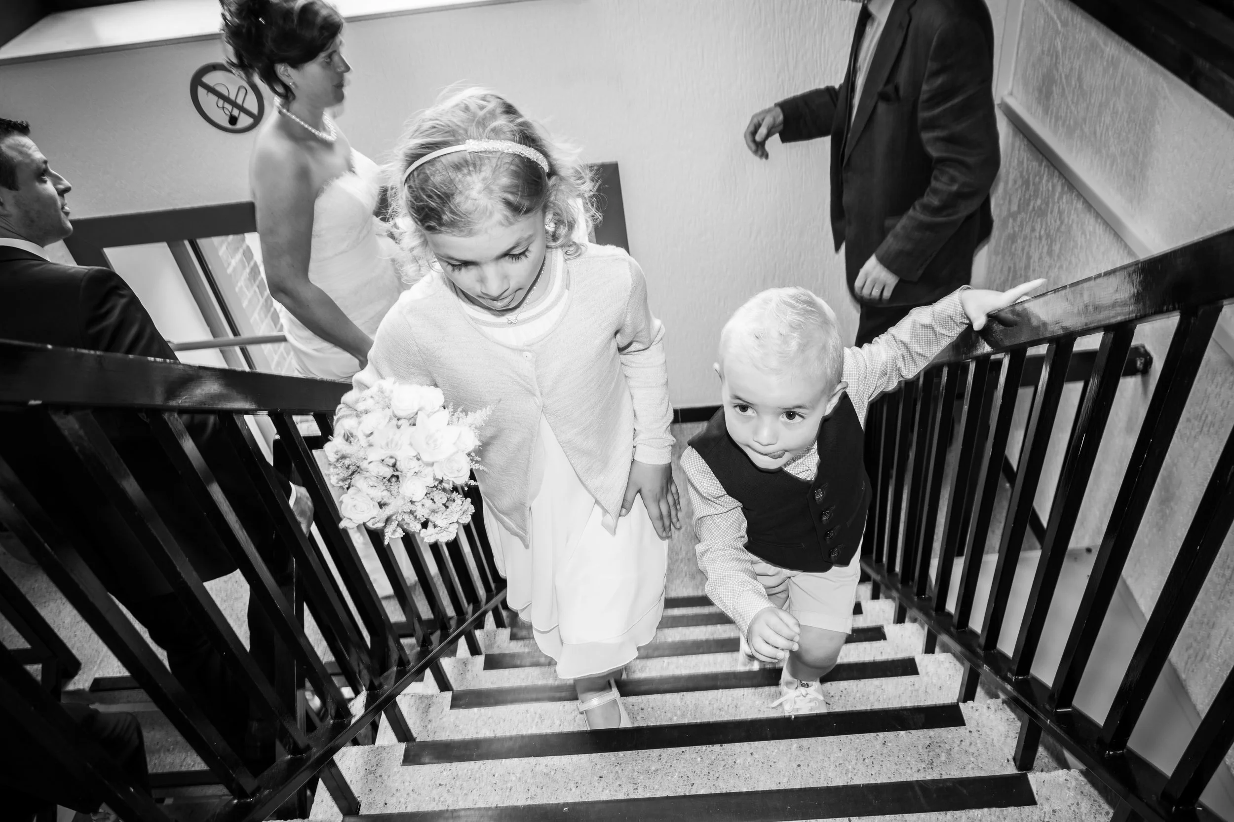 Twee jonge kinderen, een meisje en een jongen, lopen trap op tijdens een feest of bruiloft. Het meisje houdt een bloemenboeket vast.