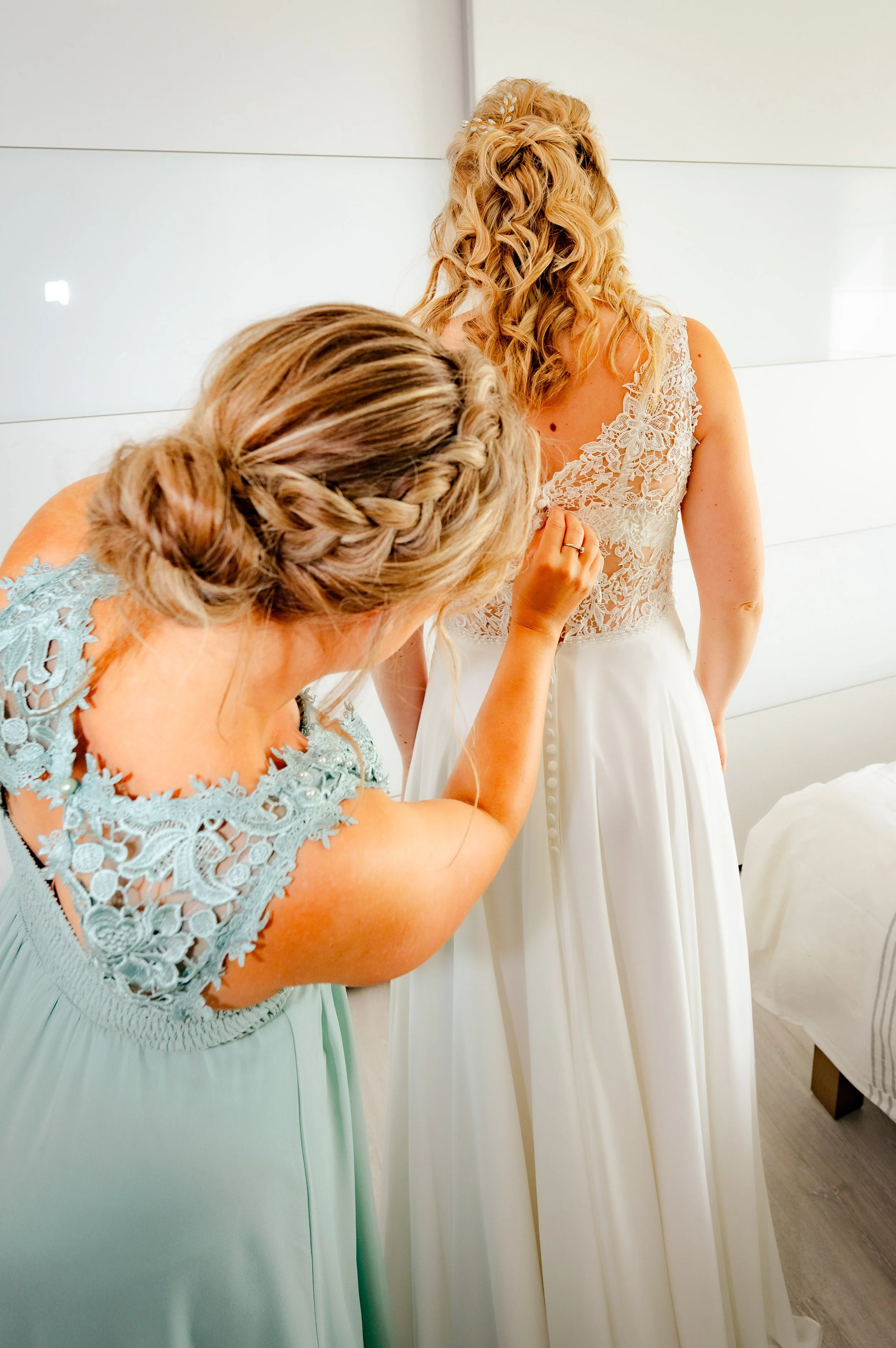 Twee vrouwen bereiden zich voor op een bruiloft; één vrouw helpt de bruid met haar jurk.
