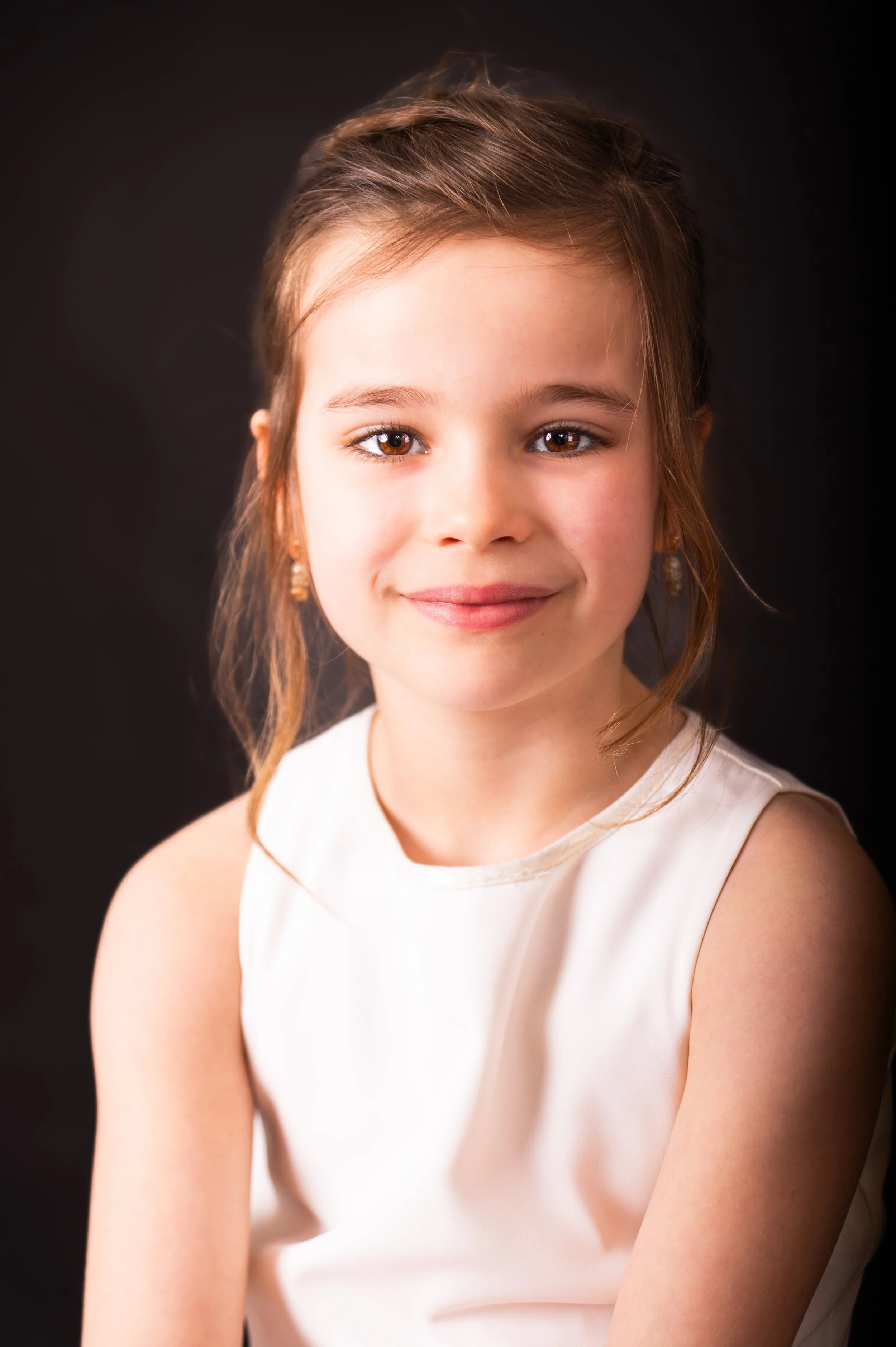 Portret van een lachende jonge vrouw met bruin haar, witte jurk en een donkere achtergrond.