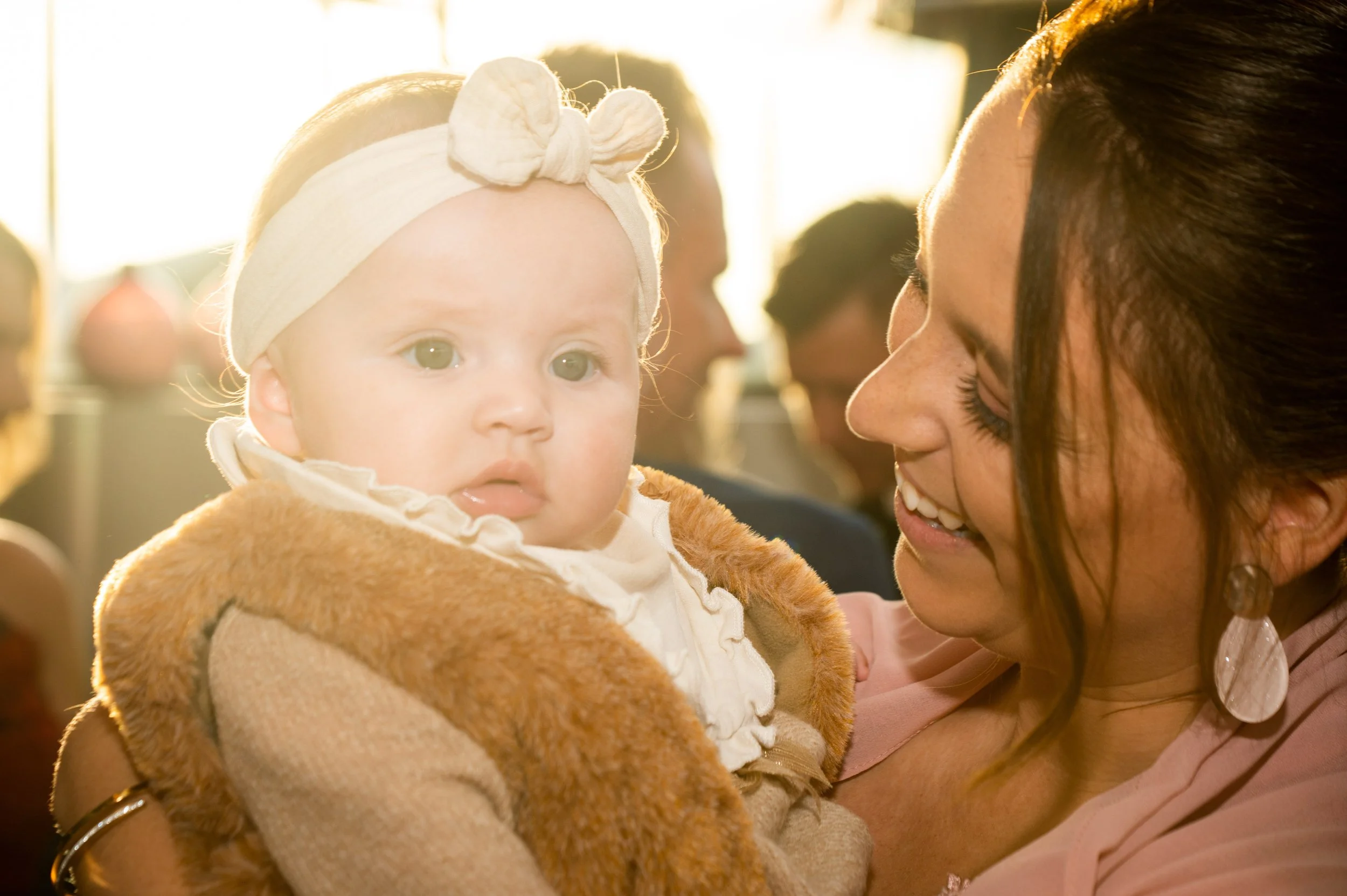 Een vrouw houdt een baby vast tijdens een zonnige gelegenheid, met andere mensen op de achtergrond.