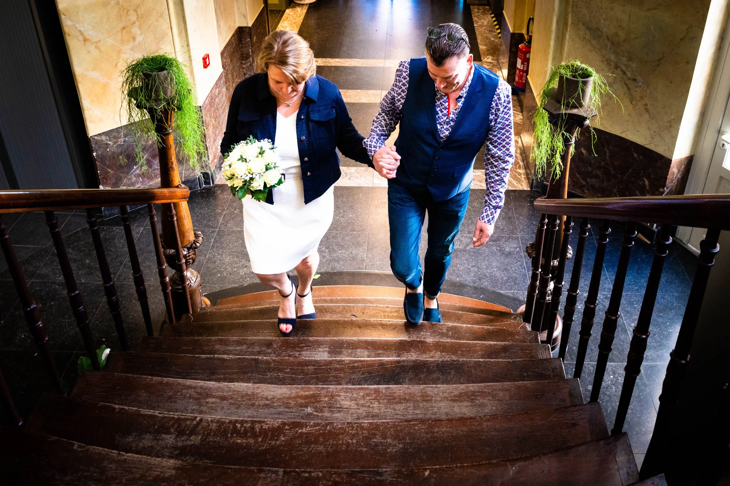 Een man en een vrouw wandelen samen op een trap in een gebouw. Ze houden elkaars hand vast en de vrouw houdt een bloemenboeket in haar linkerhand. Ze dragen nette kleding en lijken feestelijk gekleed.