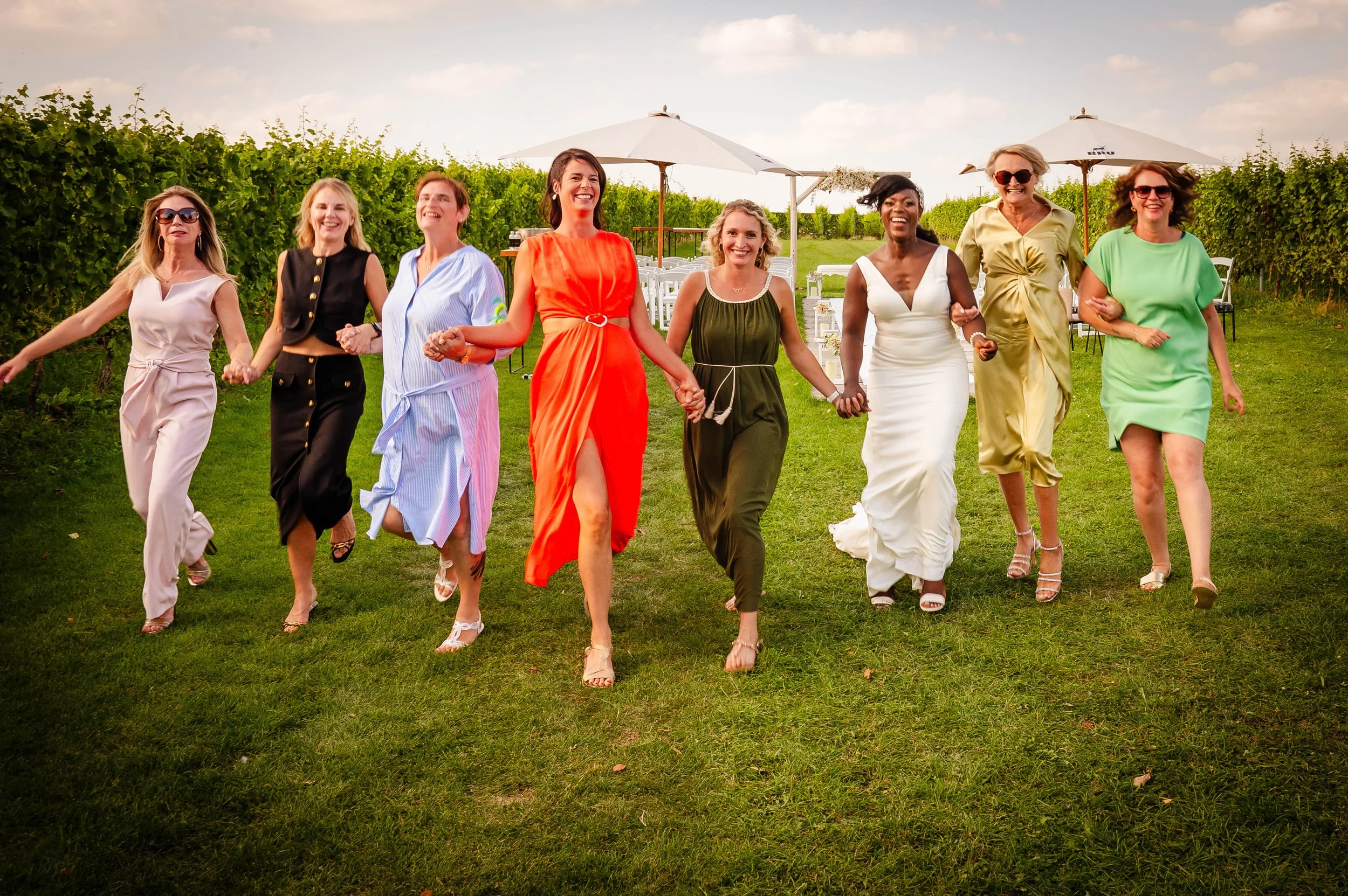 Twaalf vrouwen lopen samen hand in hand over een grasveld, lachend en plezier hebbend, op een zonnige dag met een tuin met wijnranken en picknicktafels met parasols op de achtergrond.