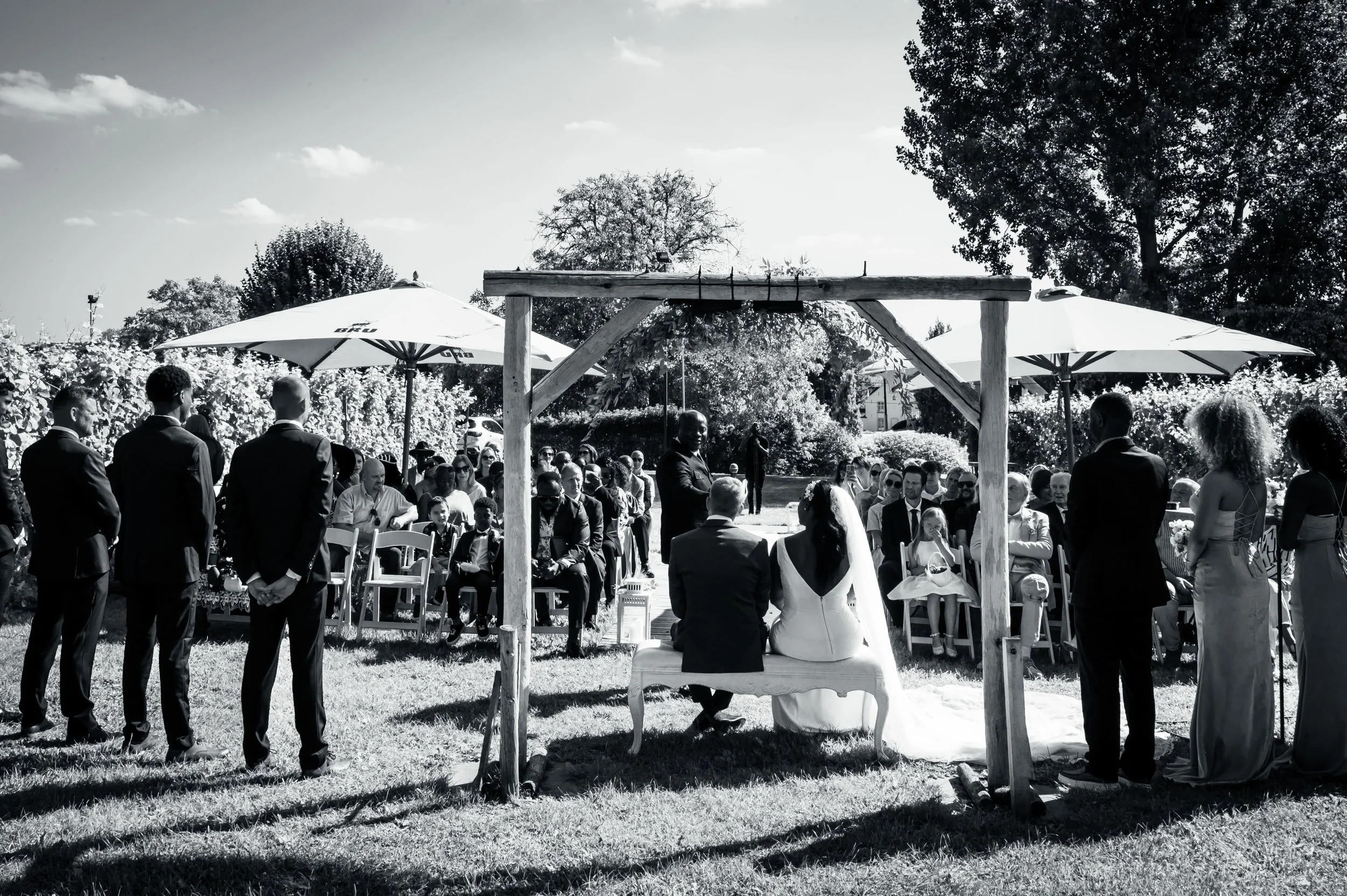 Gehuwde paar zit onder een houten boog tijdens een buitenbruiloft, omringd door gasten onder parasols op een grasveld met bomen en huizen op de achtergrond, in zwart-wit