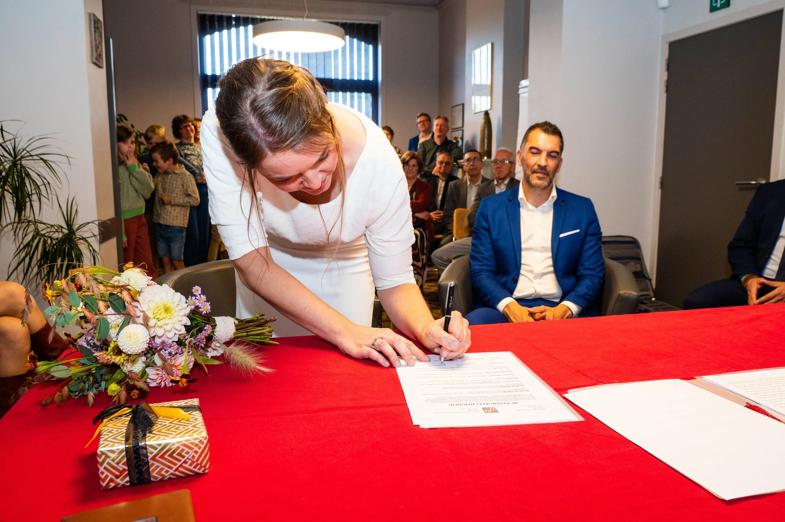 Vrouw ondertekent document tijdens een officiële ceremonie, met een bouquet bloemen en cadeau op tafel, op de achtergrond aanwezigen en een plafondlamp.