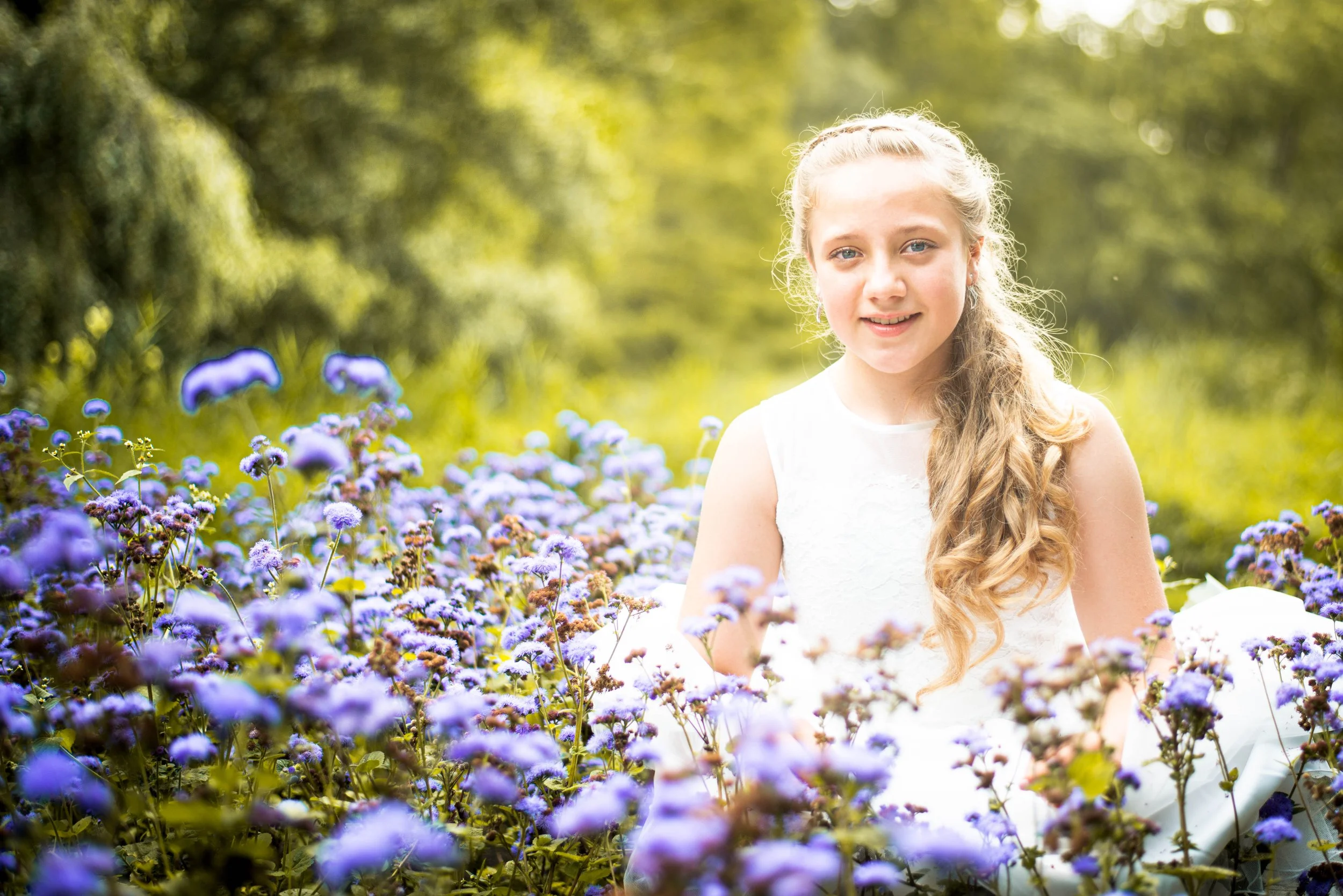 Een jonge vrouw in een witte jurk zittend in een veld met blauwe bloemen, met groen bos op de achtergrond.
